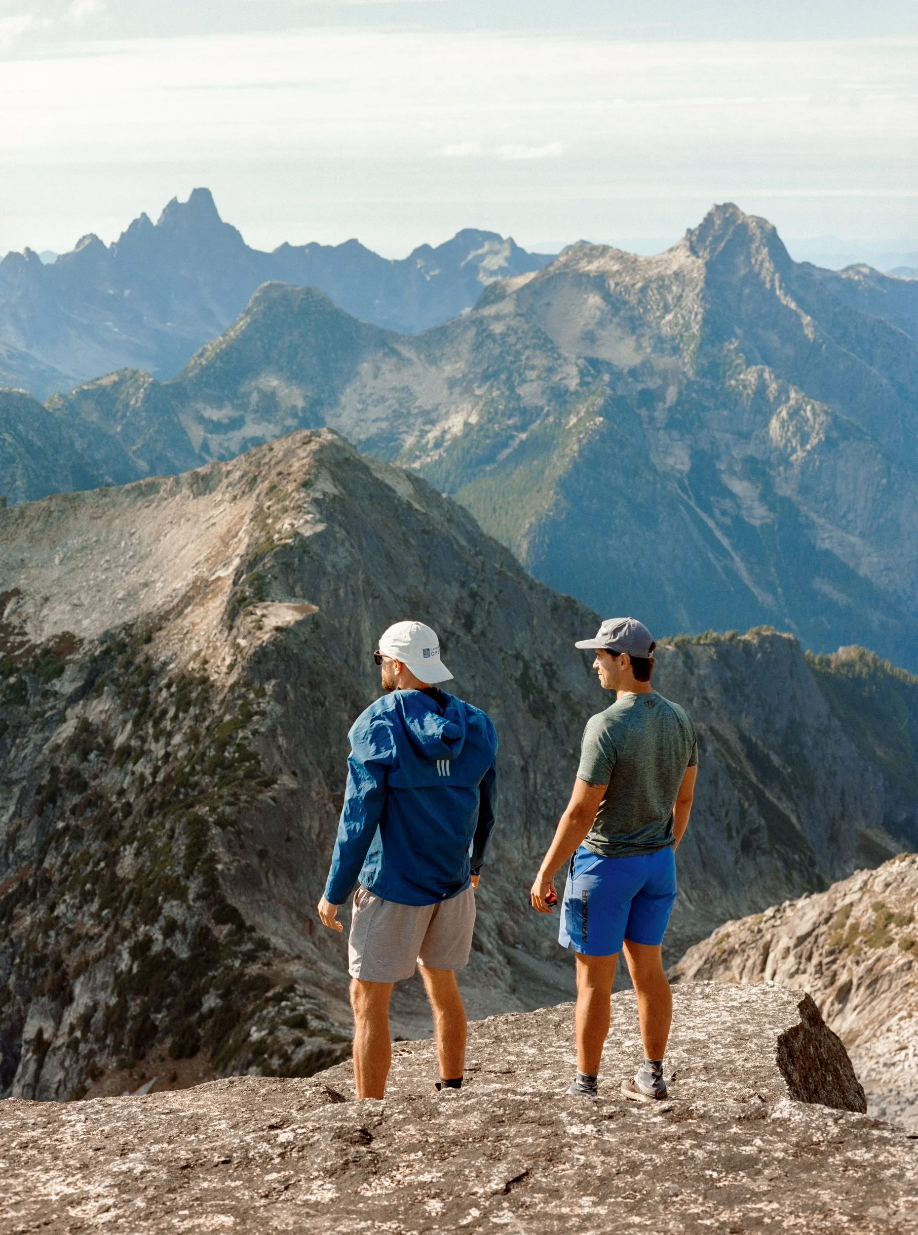 Two hikers standing on rocky terrain overlooking a mountain range with rugged peaks and deep valleys.