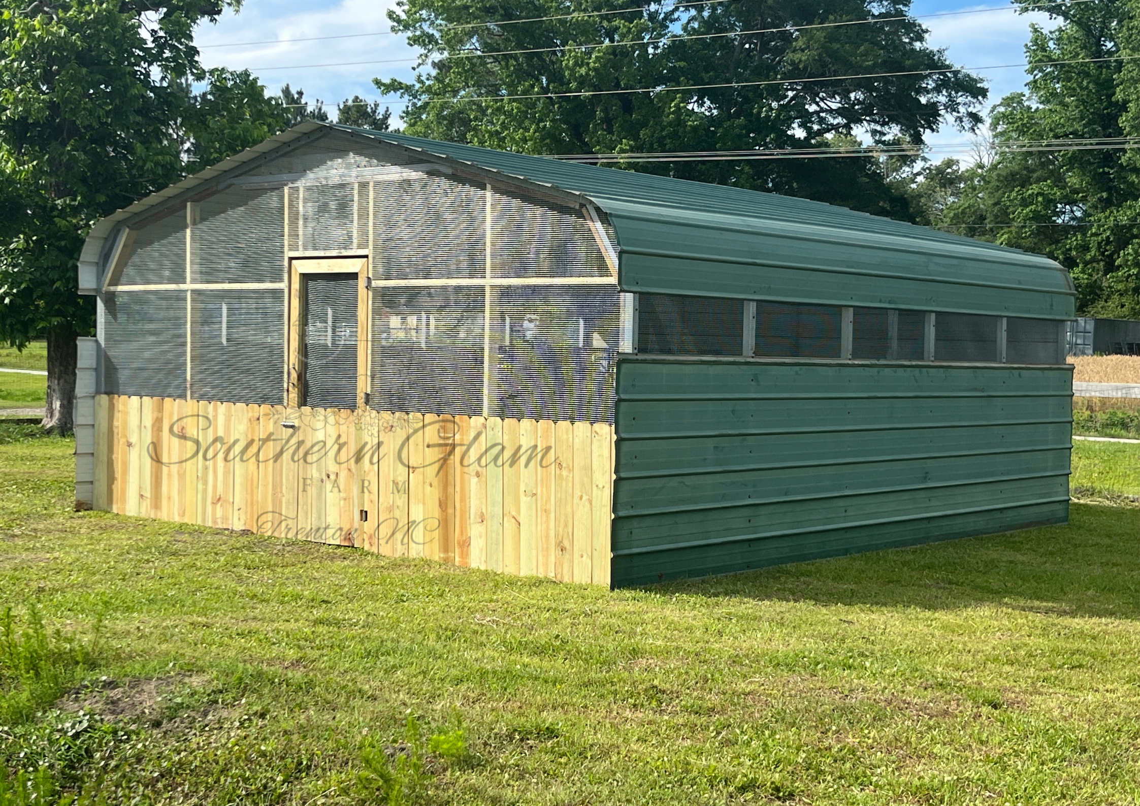 A metal chicken coop made from carport. The structure is situated on a grassy area with trees and power lines in the background.