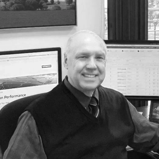 A smiling man with short light hair, wearing a dark vest over a shirt and tie, sitting at a desk in front of two computer monitors, in an office setting.