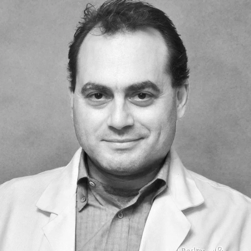 Black and white photo of a man with dark hair, wearing a collared shirt and a light-colored blazer, smiling at the camera against a plain wall background.
