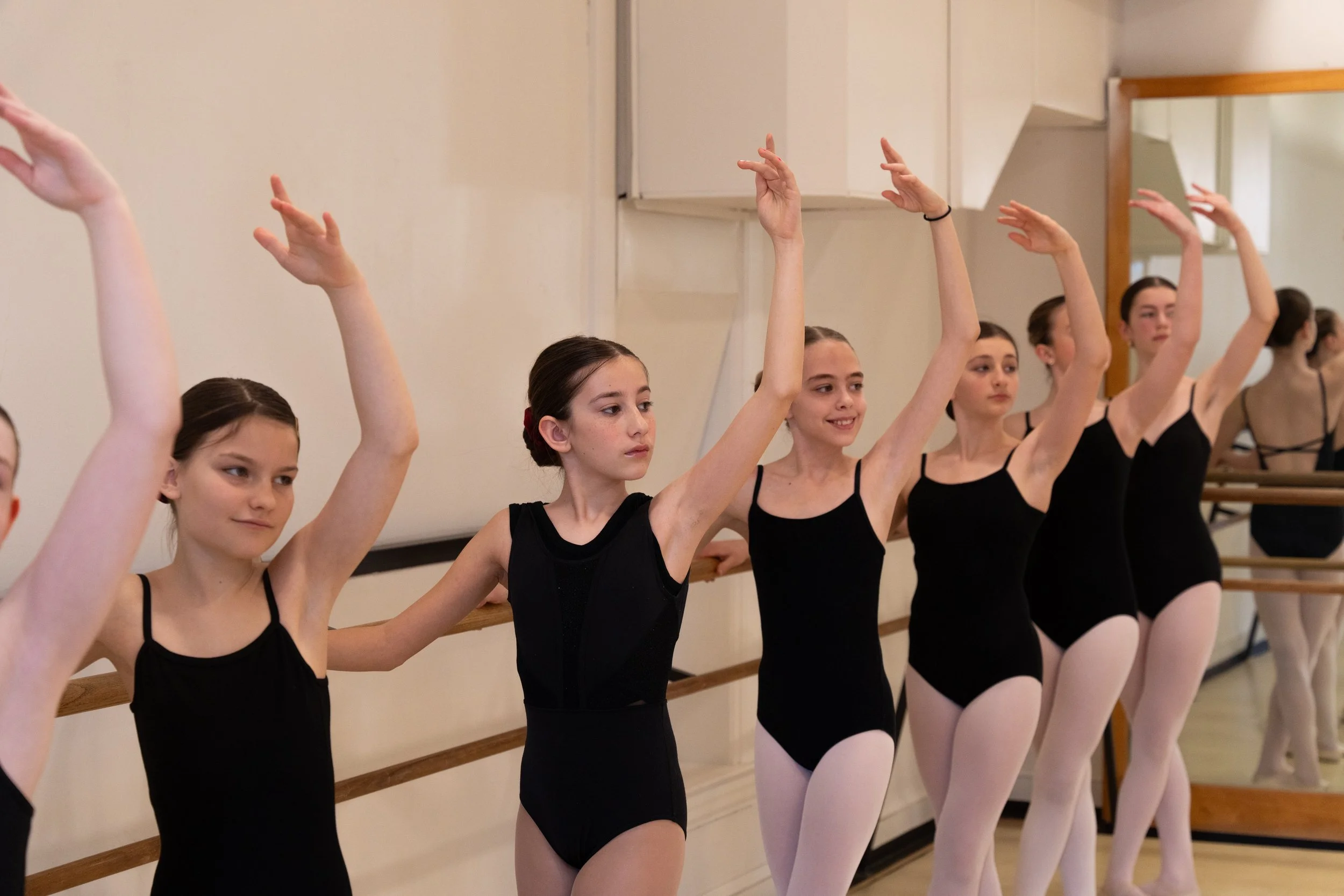 Ballet students in black leotards and pink tights practicing at a barre in a dance studio.