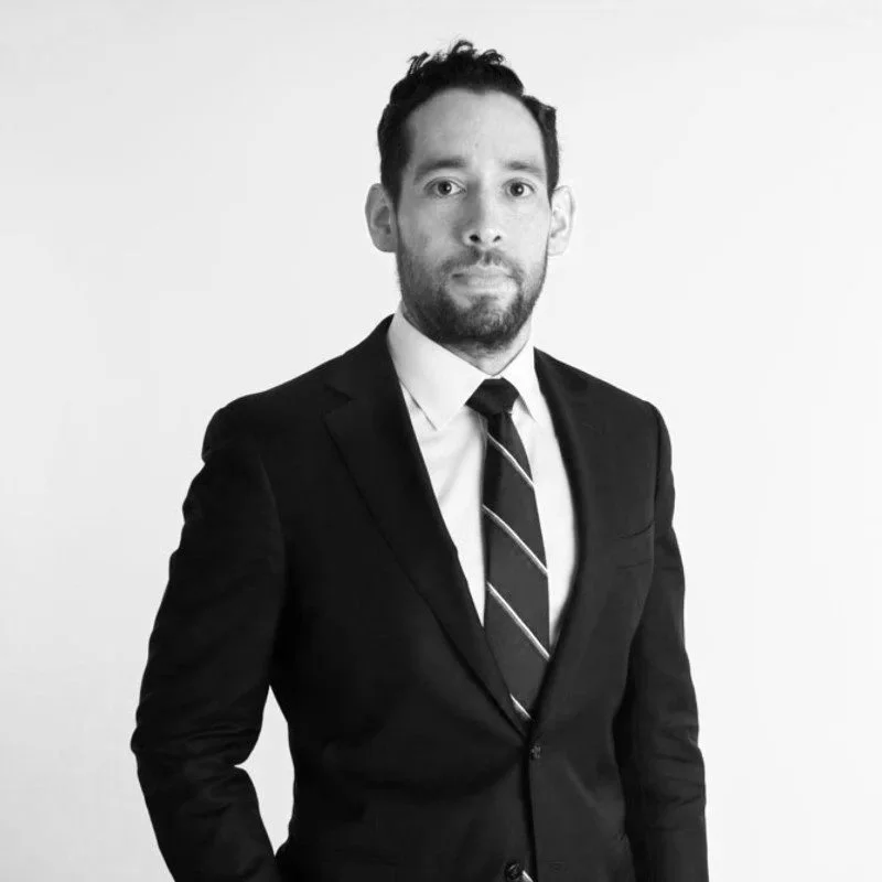 A man in a black suit, white shirt, and striped tie stands against a plain light background, looking directly at the camera.