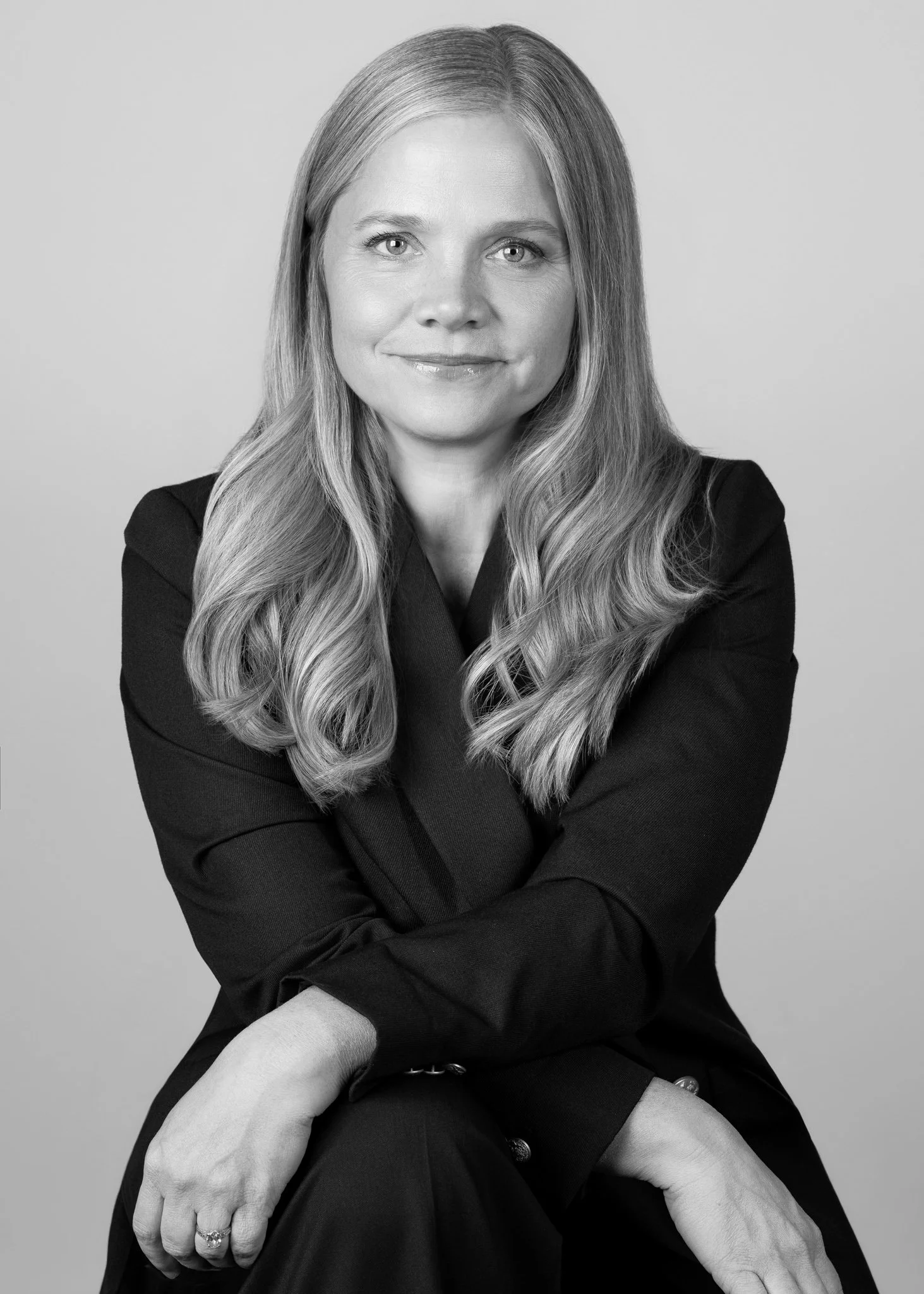 Black and white portrait of a woman with long wavy hair, wearing a black blazer, sitting with her arms crossed, smiling softly.