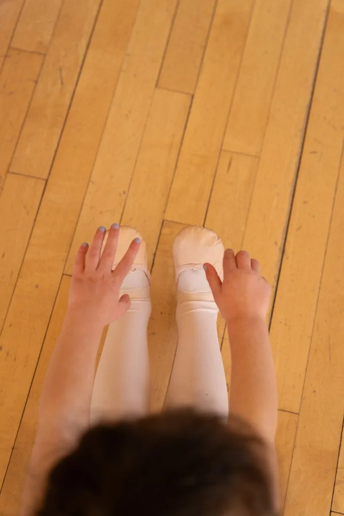 Child standing on a wooden floor wearing ballet slippers, with hands touching the slippers.