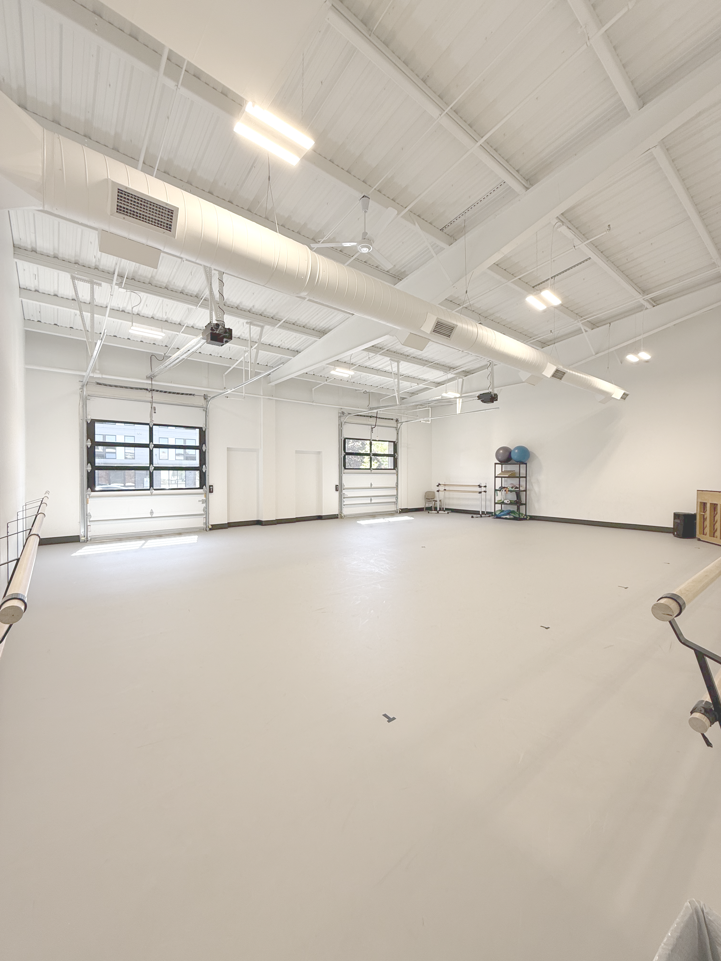 Wide, empty fitness studio with light-colored floor, high ceiling with white pipes and beams, windows on one wall, ballet barres, and fitness balls on a shelf.