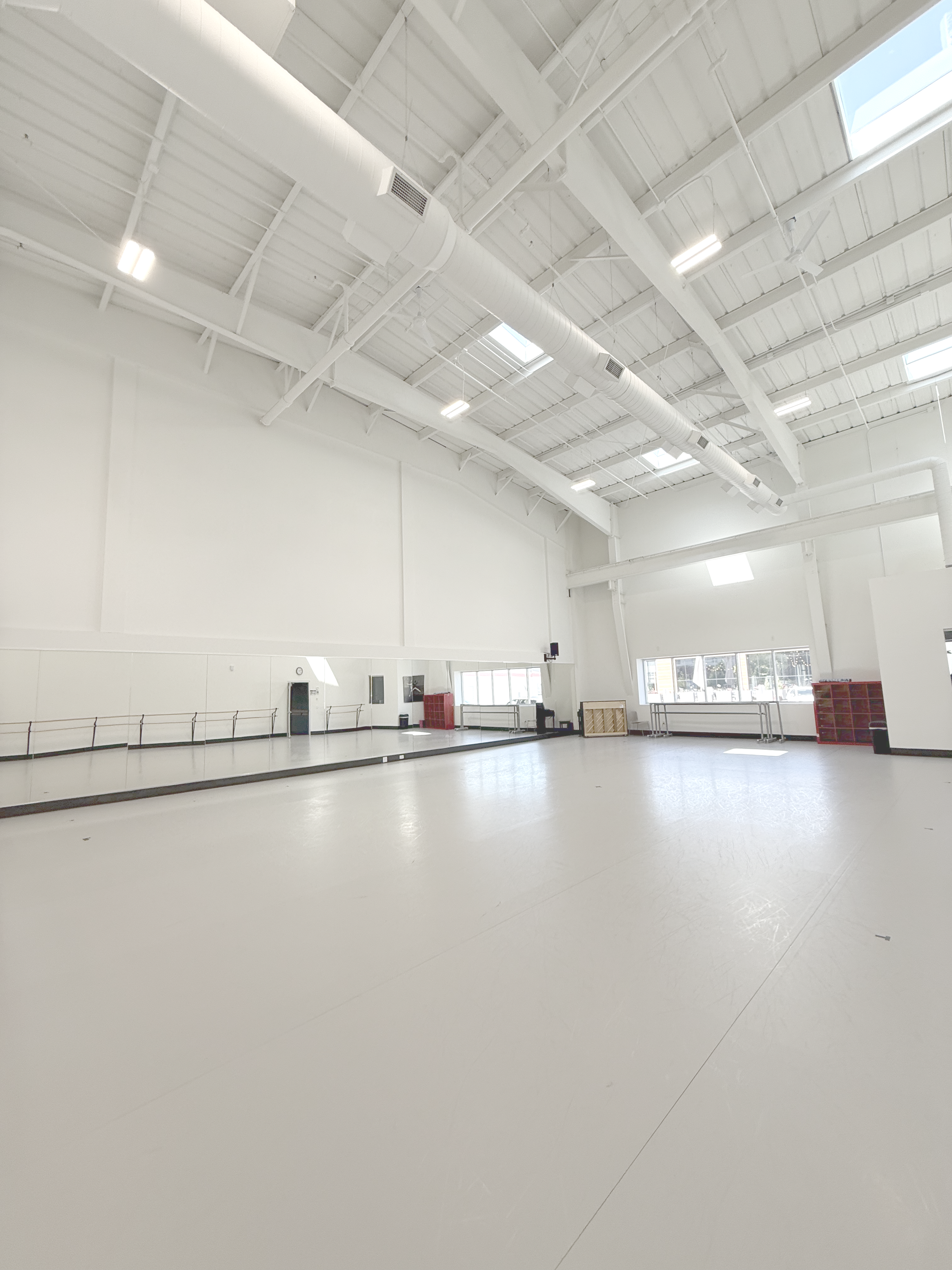 A spacious dance studio with white walls, high ceiling with skylights, ballet barres, and mirrors, equipped for dance practice.