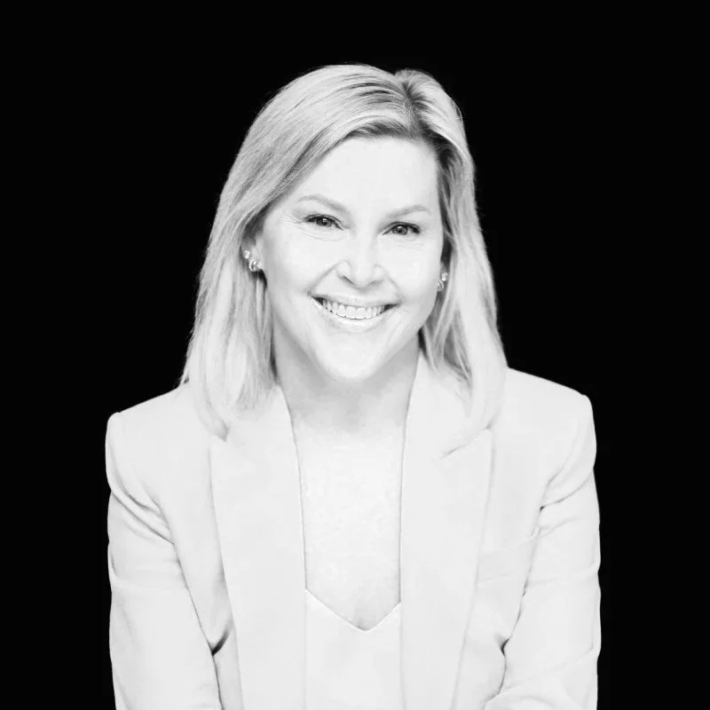 A woman with shoulder-length blonde hair smiling, wearing a light-colored blazer, against a black background.