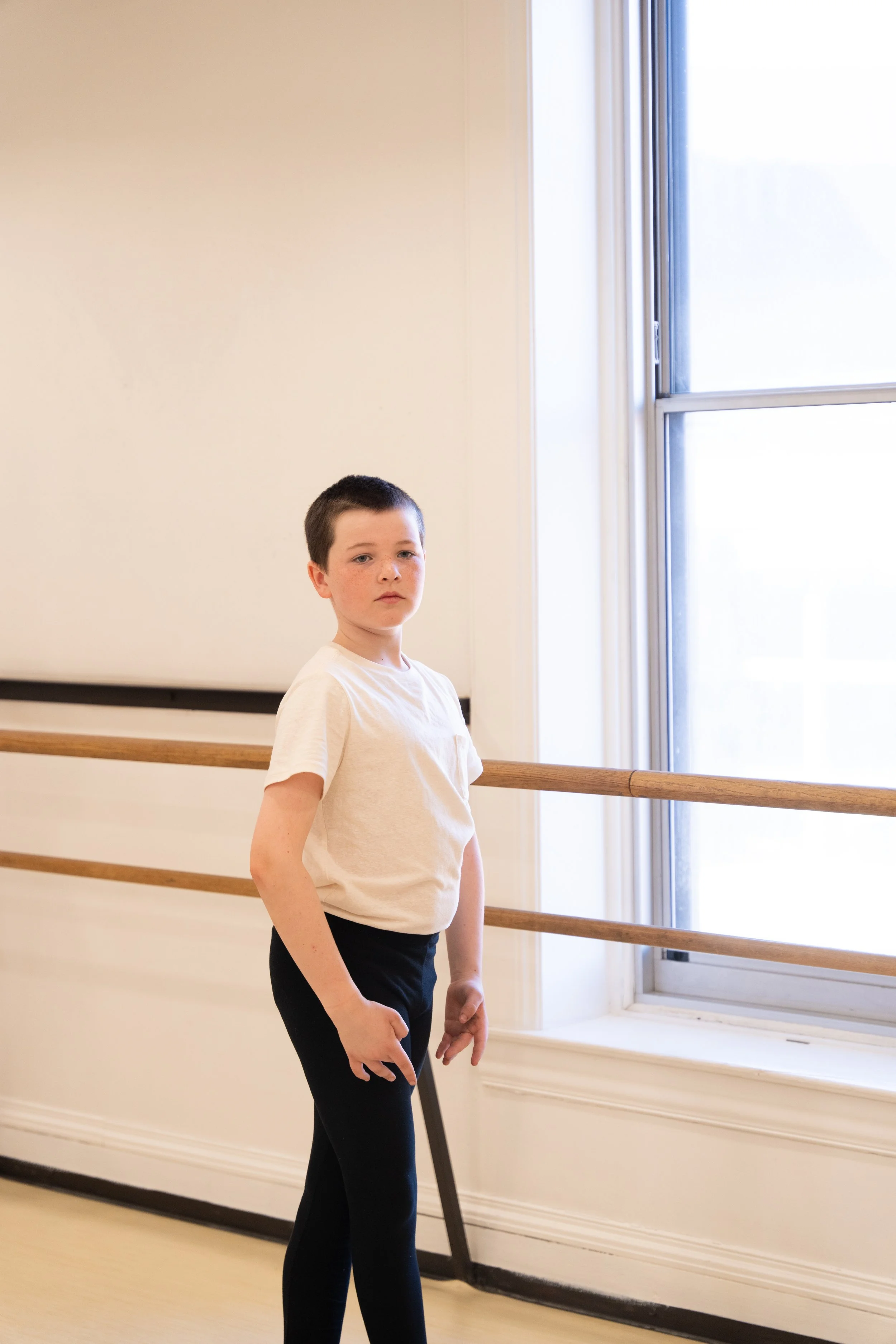 A young boy with short dark hair, wearing a white t-shirt and black pants, standing in a dance studio next to a wooden ballet barre and a large window with frosted glass.