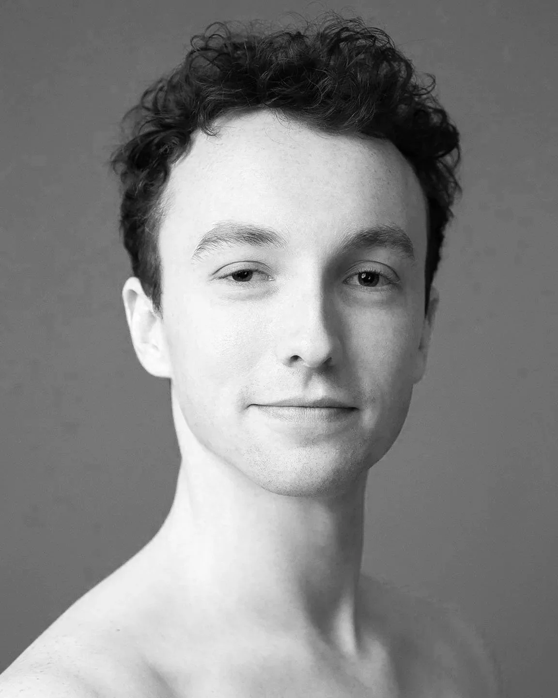 Black-and-white portrait of a young man with curly hair, slightly smiling, looking directly at the camera with a neutral background.