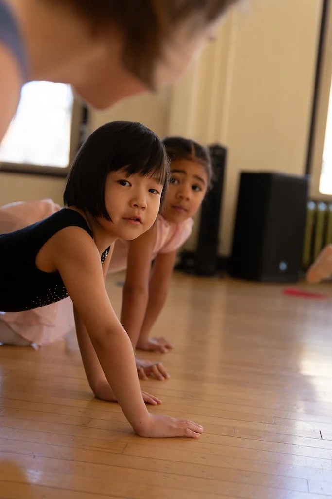 Two young girls and an adult woman on their hands and knees on a wooden floor, possibly doing yoga or exercise, in a room with a speaker and windows.