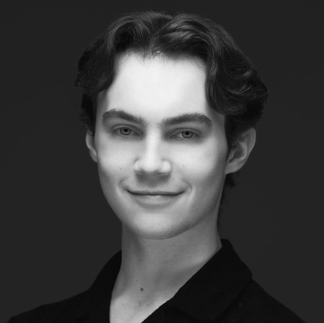 Black and white portrait of a young man with wavy dark hair, wearing a dark collared shirt, smiling softly against a dark background.