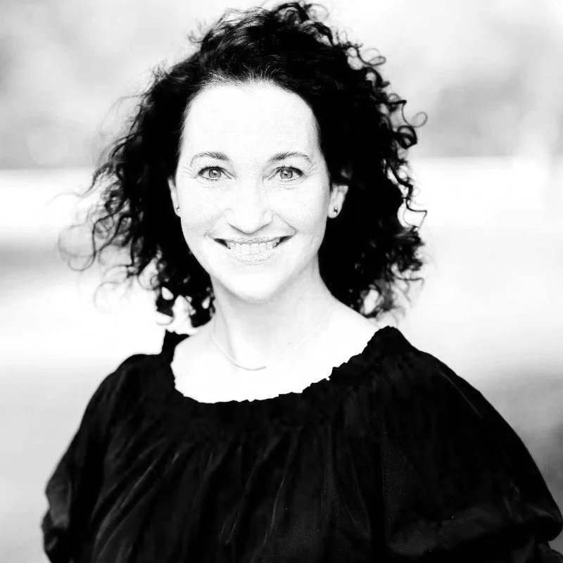 A woman with curly hair smiling at the camera in a black top outdoors.