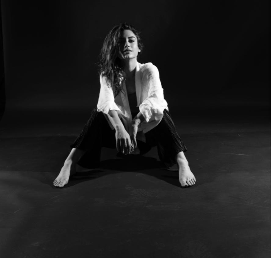 Black and white photo of a woman sitting on the floor with legs apart and hands on her ankles, wearing a light jacket and dark pants, with tousled hair and an intense expression, against a dark background.