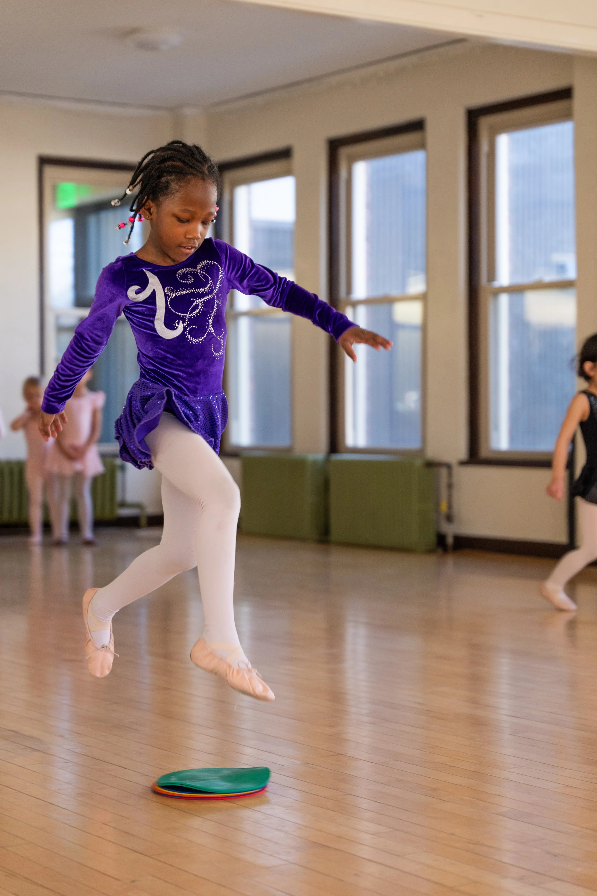 A young girl wearing a purple velvet dress with rhinestone designs, white leggings, and ballet slippers is jumping in a dance studio during a dance class. Other children are visible in the background.