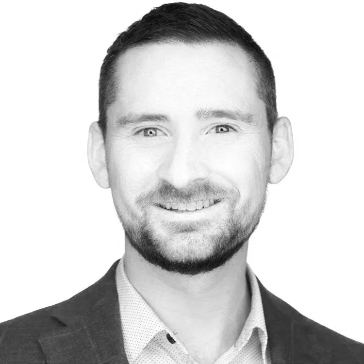 Black and white portrait of a smiling man with short dark hair and a beard, wearing a suit jacket and a collared shirt.