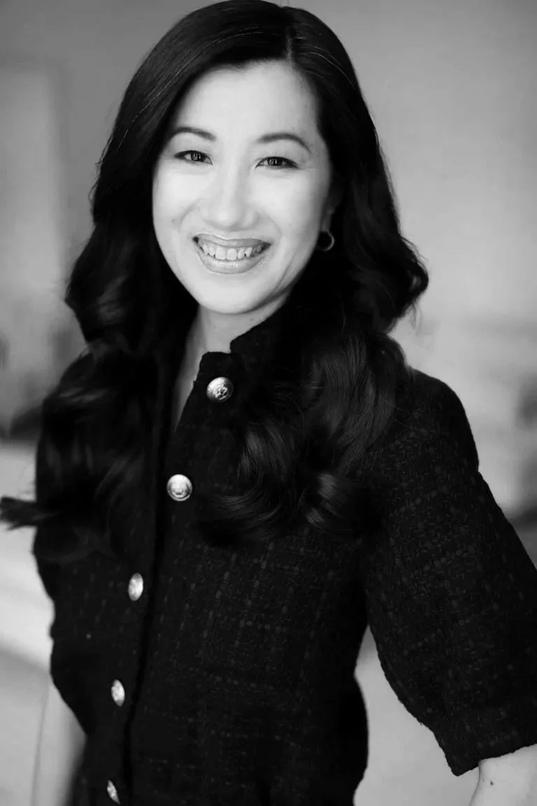 Black and white portrait of a woman with long, wavy hair smiling and wearing a dark textured jacket with metallic buttons.