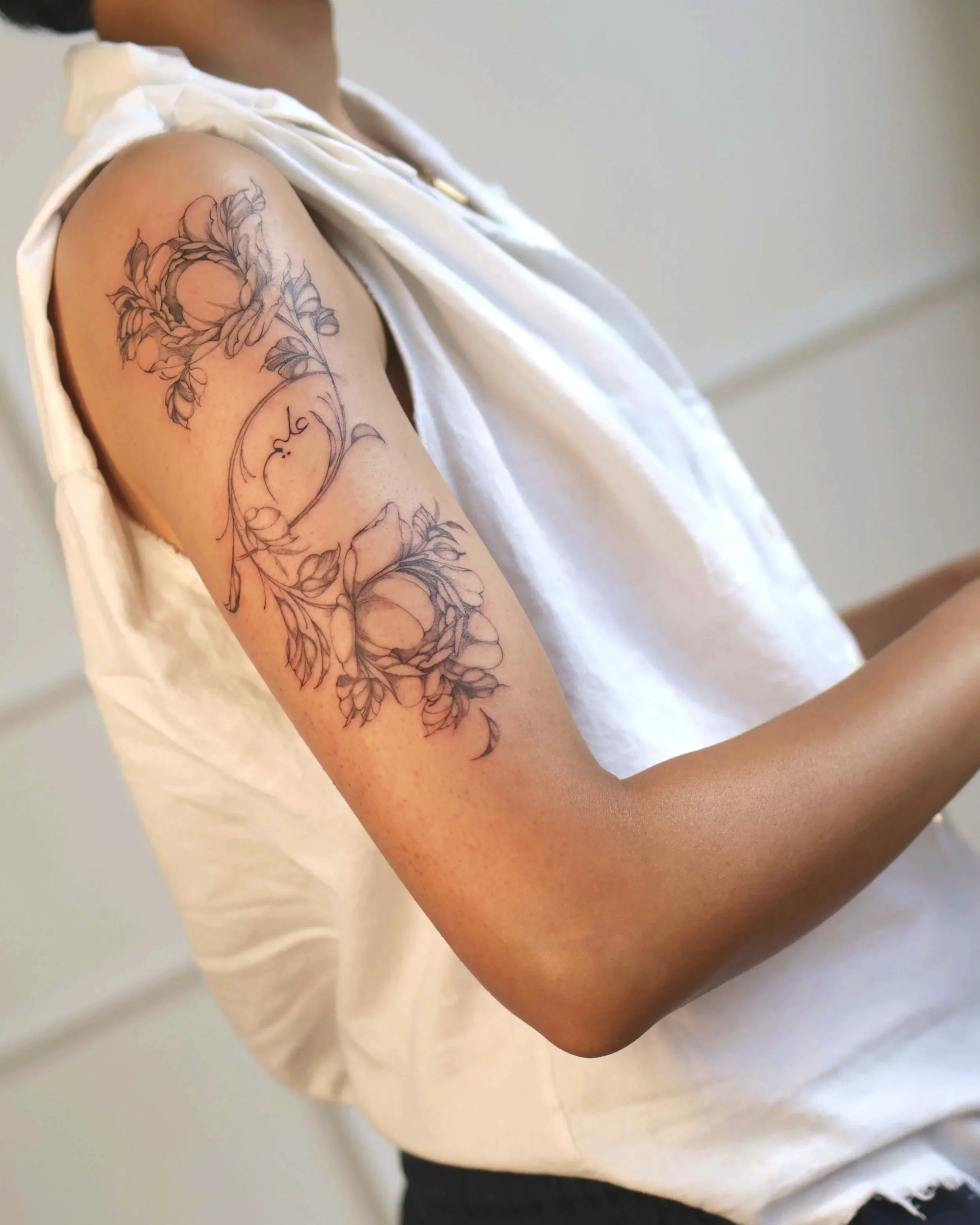 Close-up of a person's upper arm with a detailed black ink floral tattoo and a white sleeveless top.