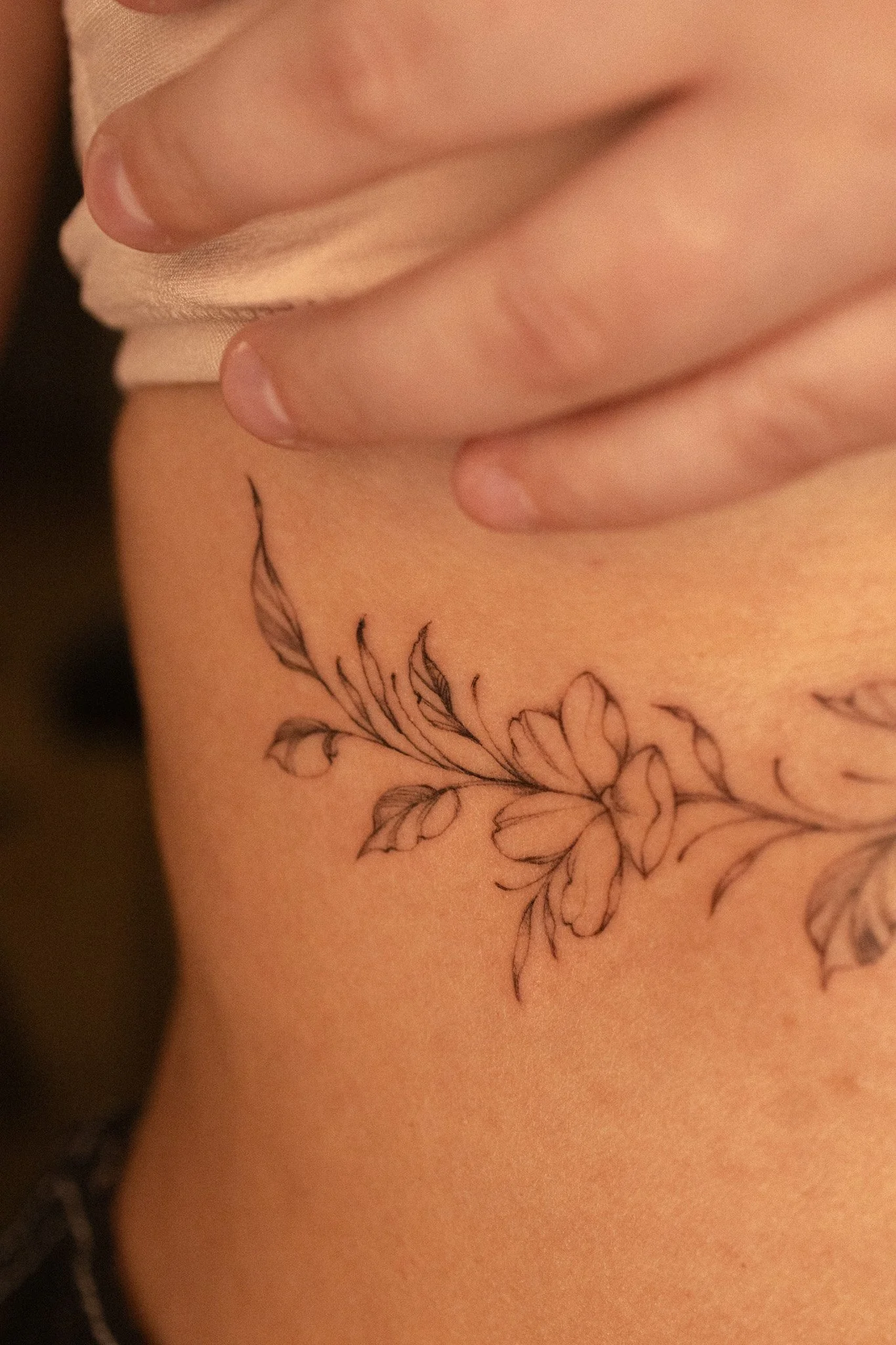 A floral tattoo on the ribs with a cherry blossom surrounded by leaves. Five petal flower, clean line work, light shading