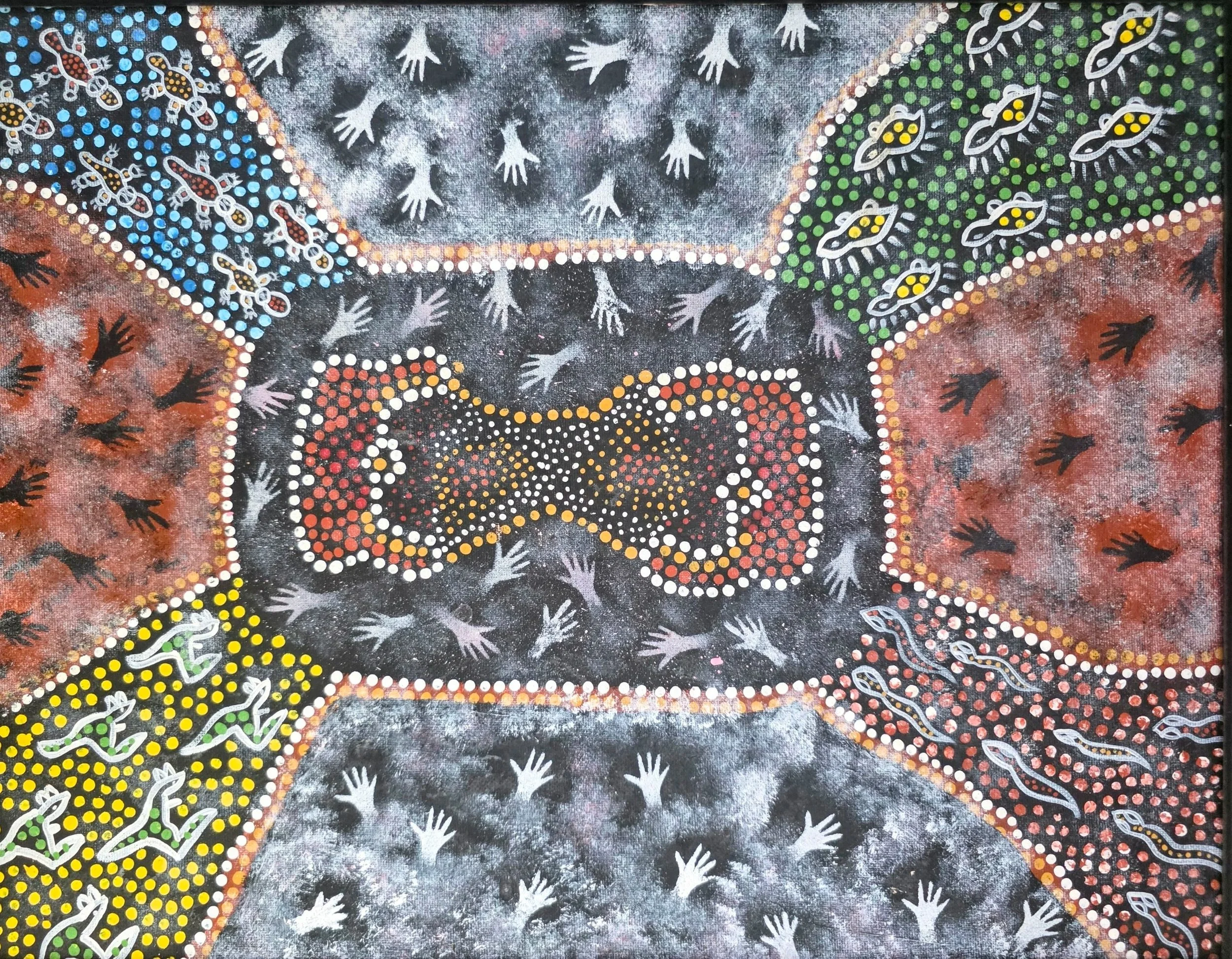 A colorful Indigenous art painting with various patterns, animal figures, and human hand prints surrounding a central design resembling an animal or symbol.