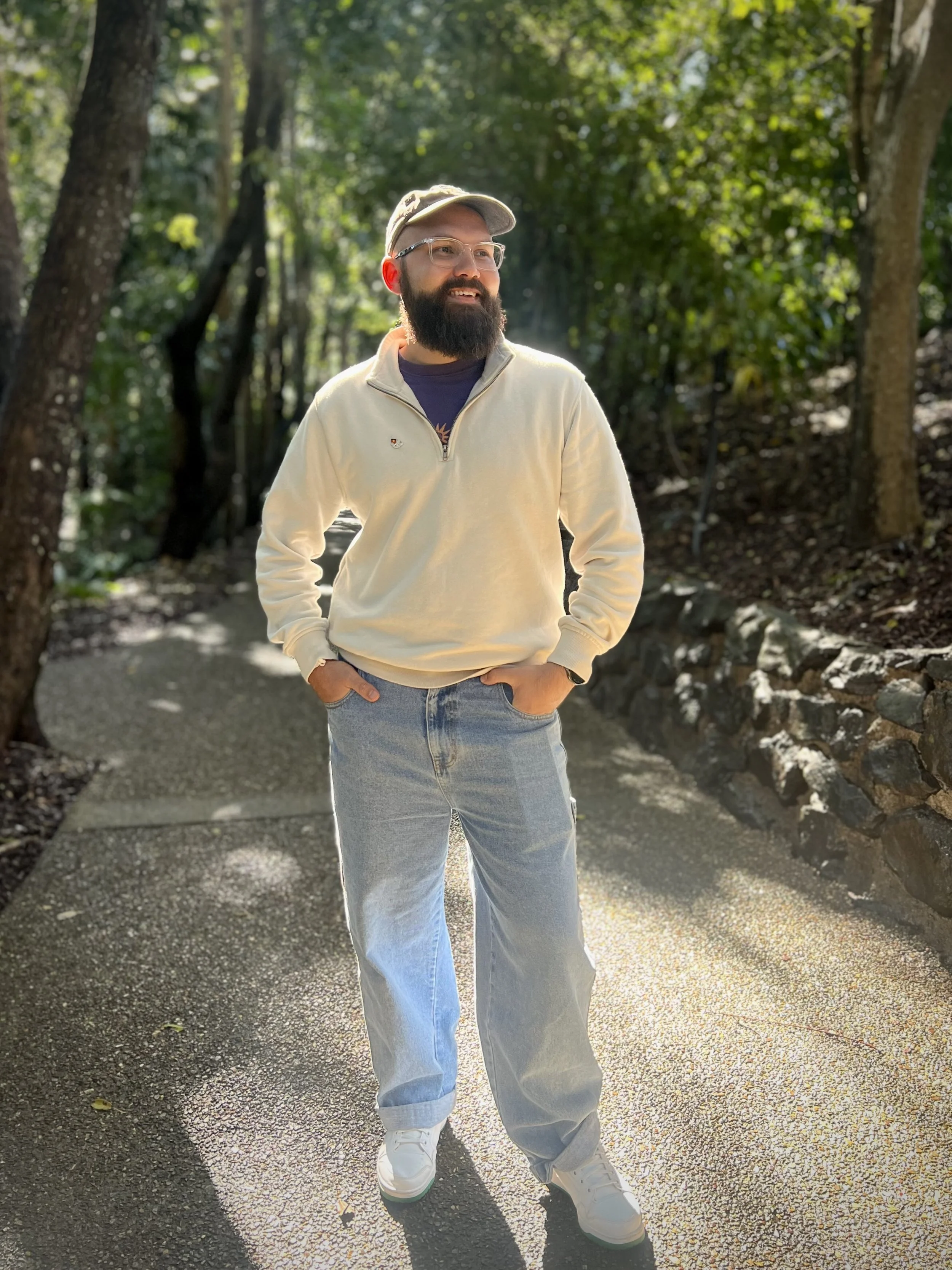 A man with a beard, wearing glasses, a beige cap, a beige quarter-zip pullover, and light blue jeans, stands on a pathway in a wooded outdoor setting, smiling and looking to the side.