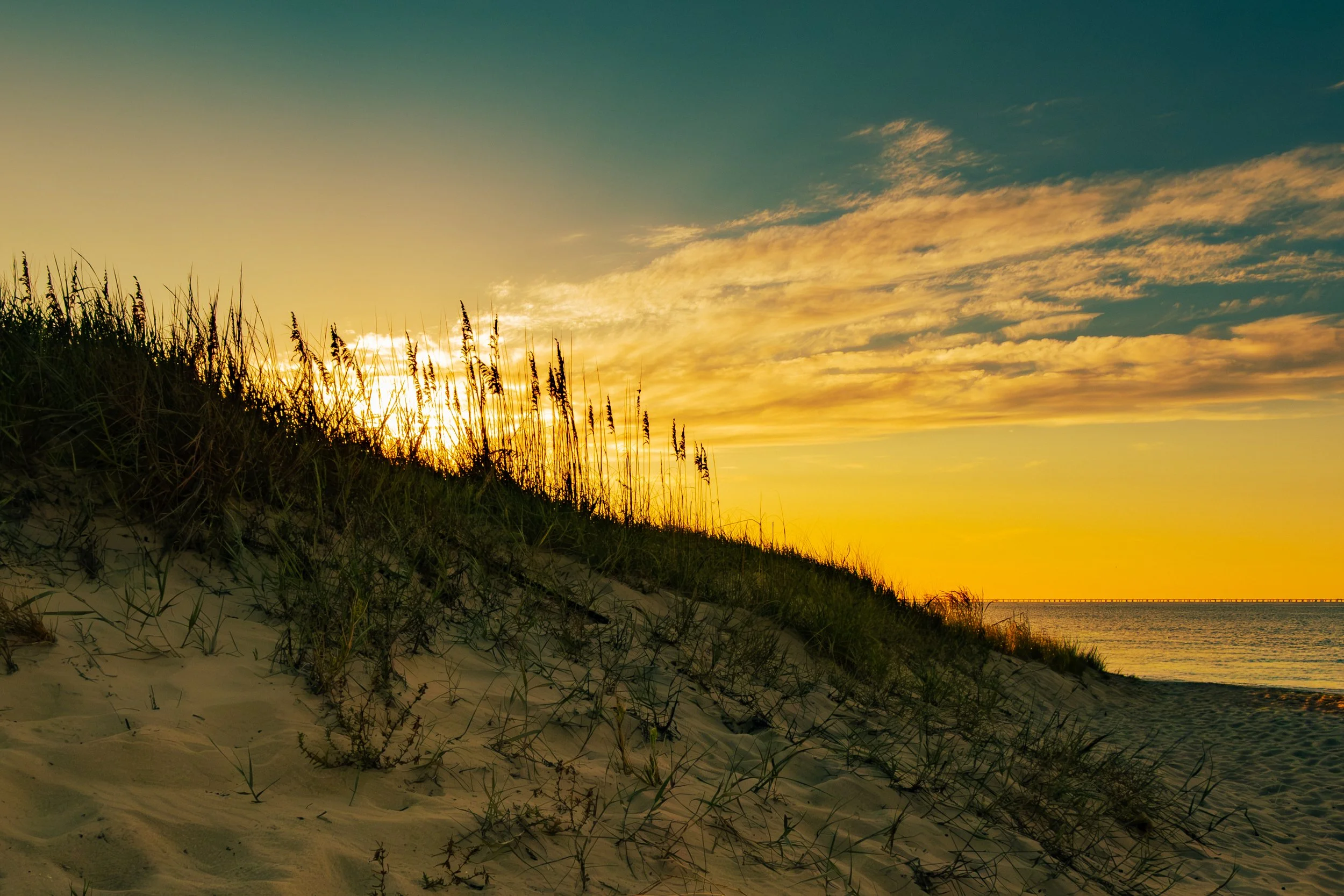 A sandy beach with grassy dunes in the foreground and the sun setting on the horizon, casting a golden glow across the sky and water, with some clouds overhead.