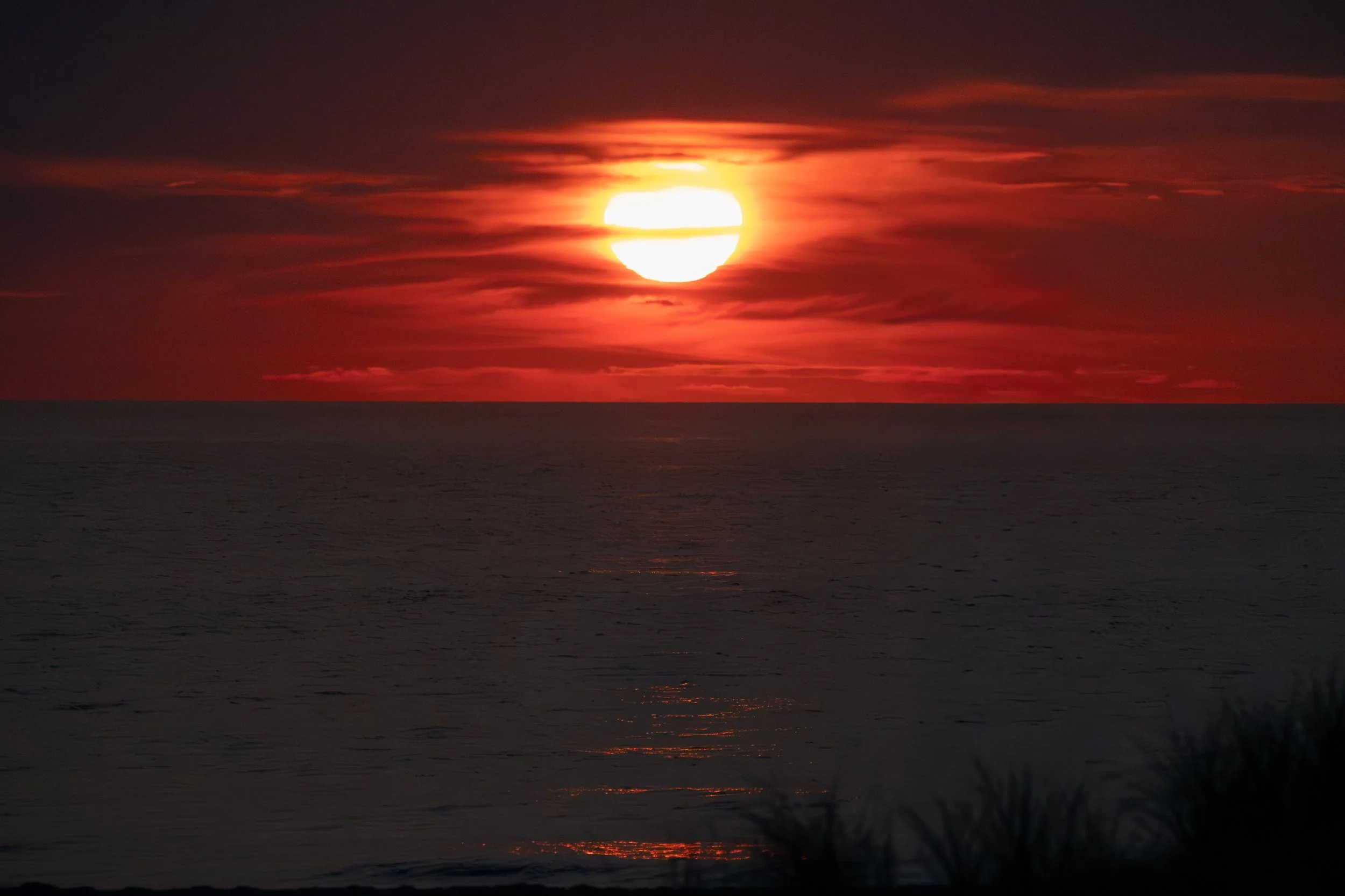 A sunset over the ocean with a large, glowing sun partially obscured by dark clouds, creating an orange and red sky reflected on the water.