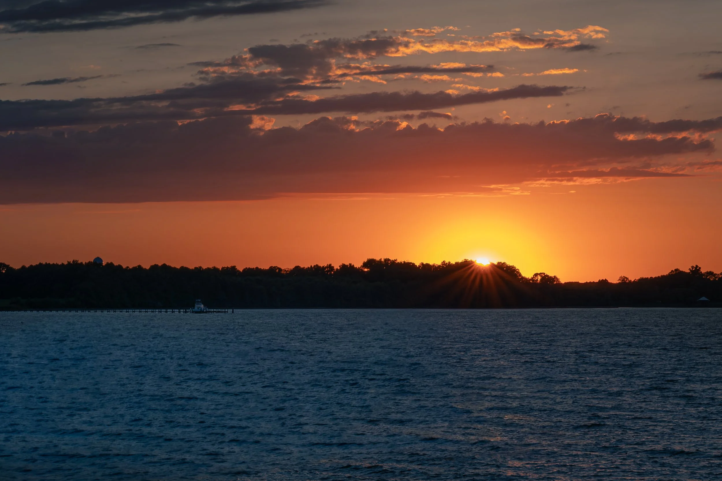 Sunset over a body of water with a distant shoreline and cloudy sky.