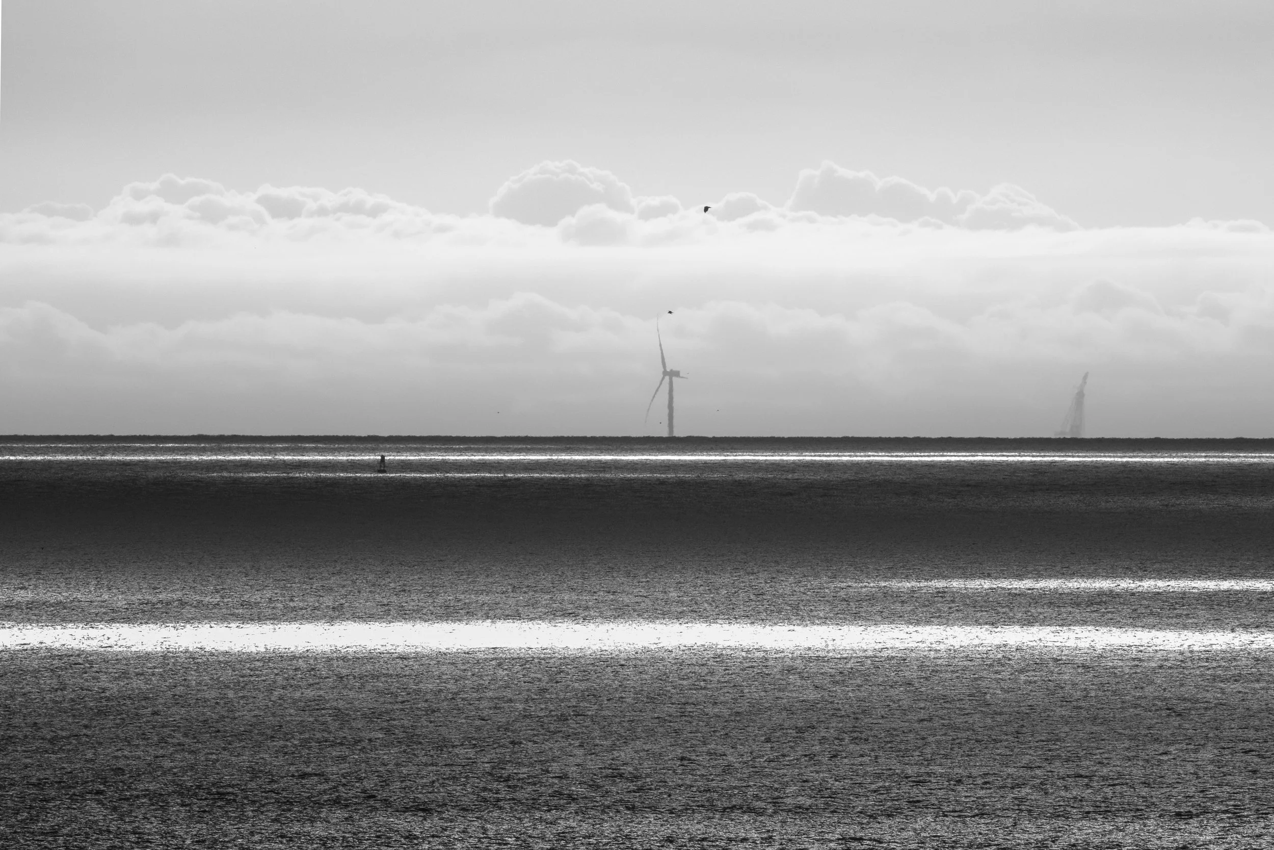 A black and white photo of a flat ocean or sea with wind turbines far in the distance and clouds in the sky.
