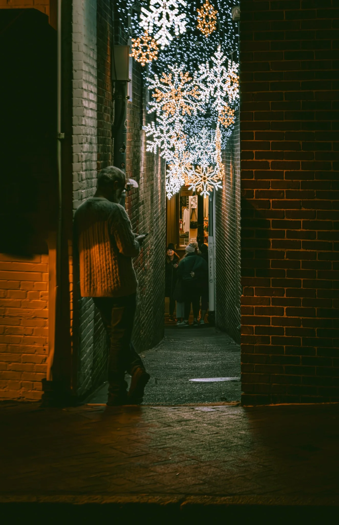 Nighttime scene of a narrow alleyway with illuminated holiday snowflake decorations hanging above. A man leans against one wall, looking at his phone, while a group of people stand further down in the alley.