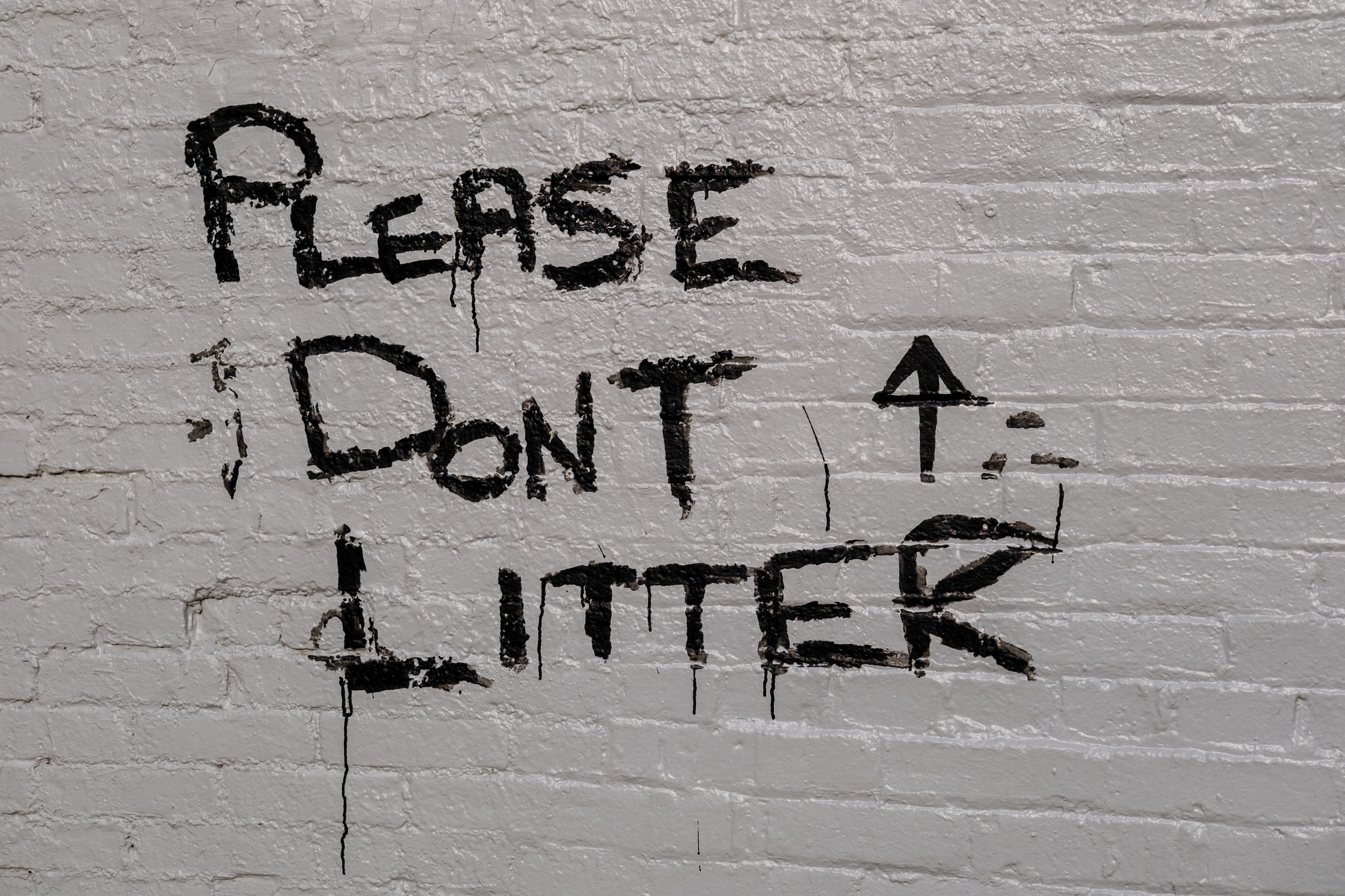 Graffiti on a white brick wall saying, 'Please don't' with an arrow pointing up and the word 'Litter' underneath.