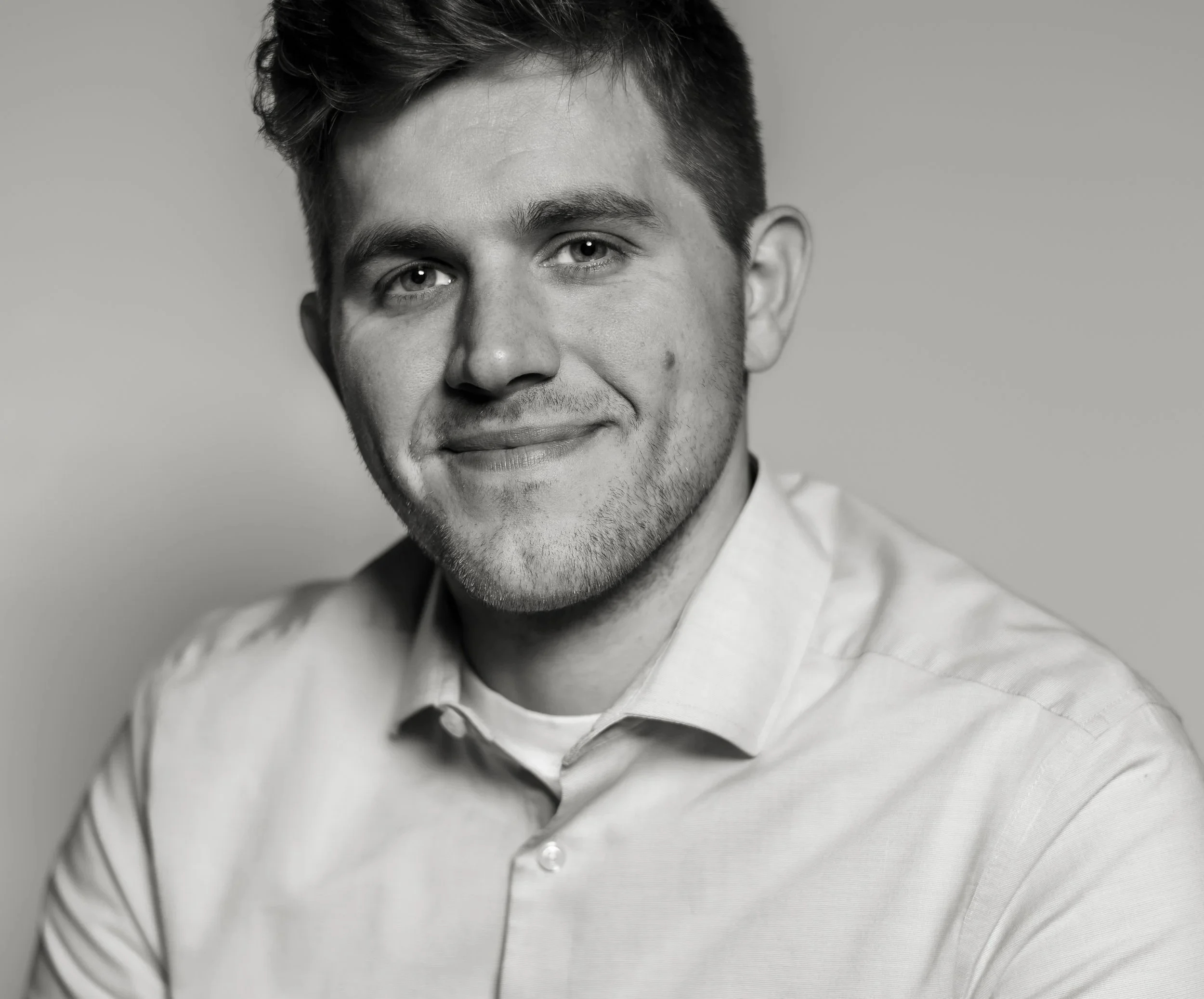 Black and white portrait of a young man with short, styled hair, wearing a light-colored button-up shirt, smiling slightly and looking directly at the camera.
