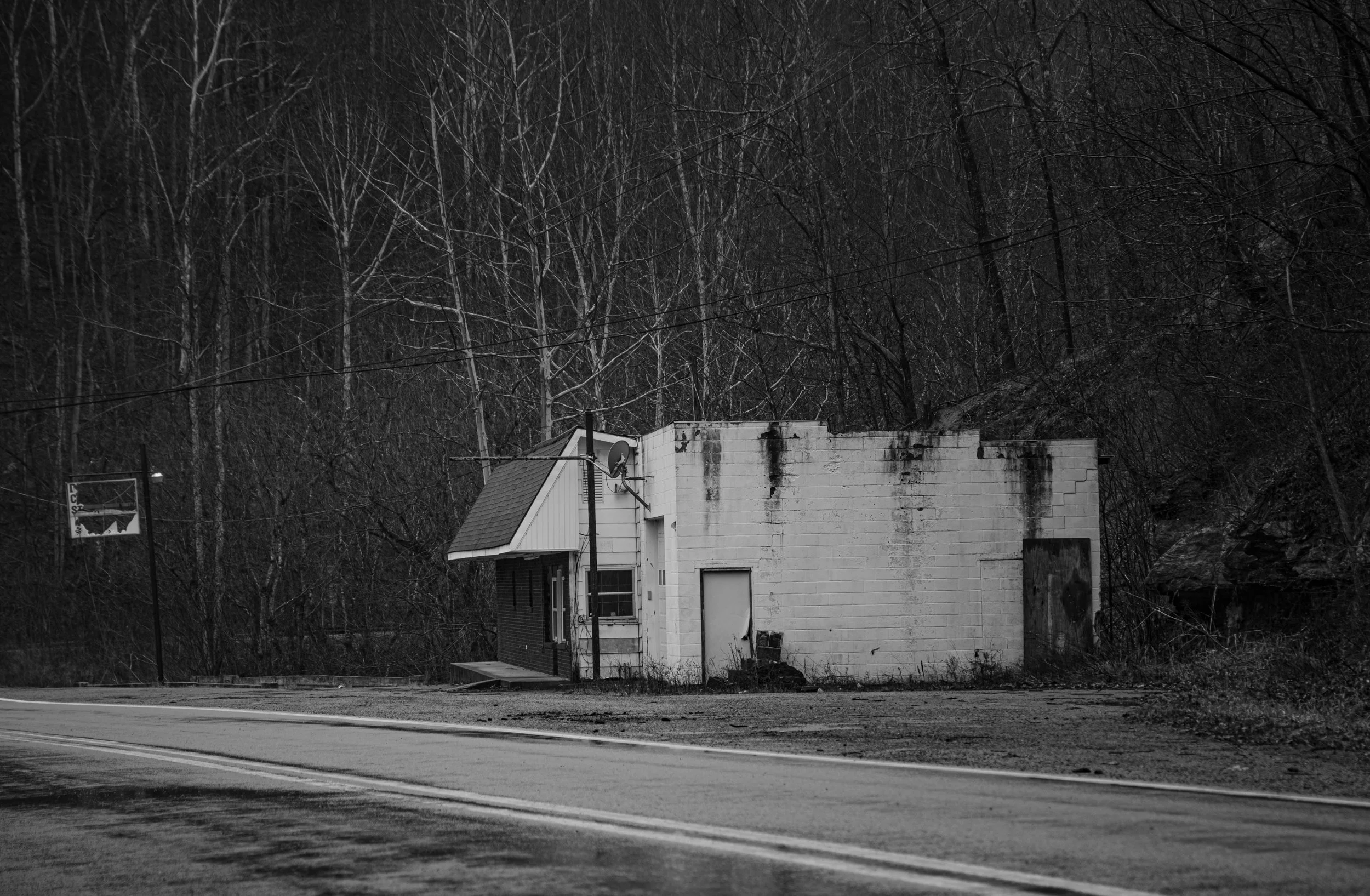 Black and white photograph of an abandoned building next to a road, with trees in the background and a partially visible electric sign to the left.