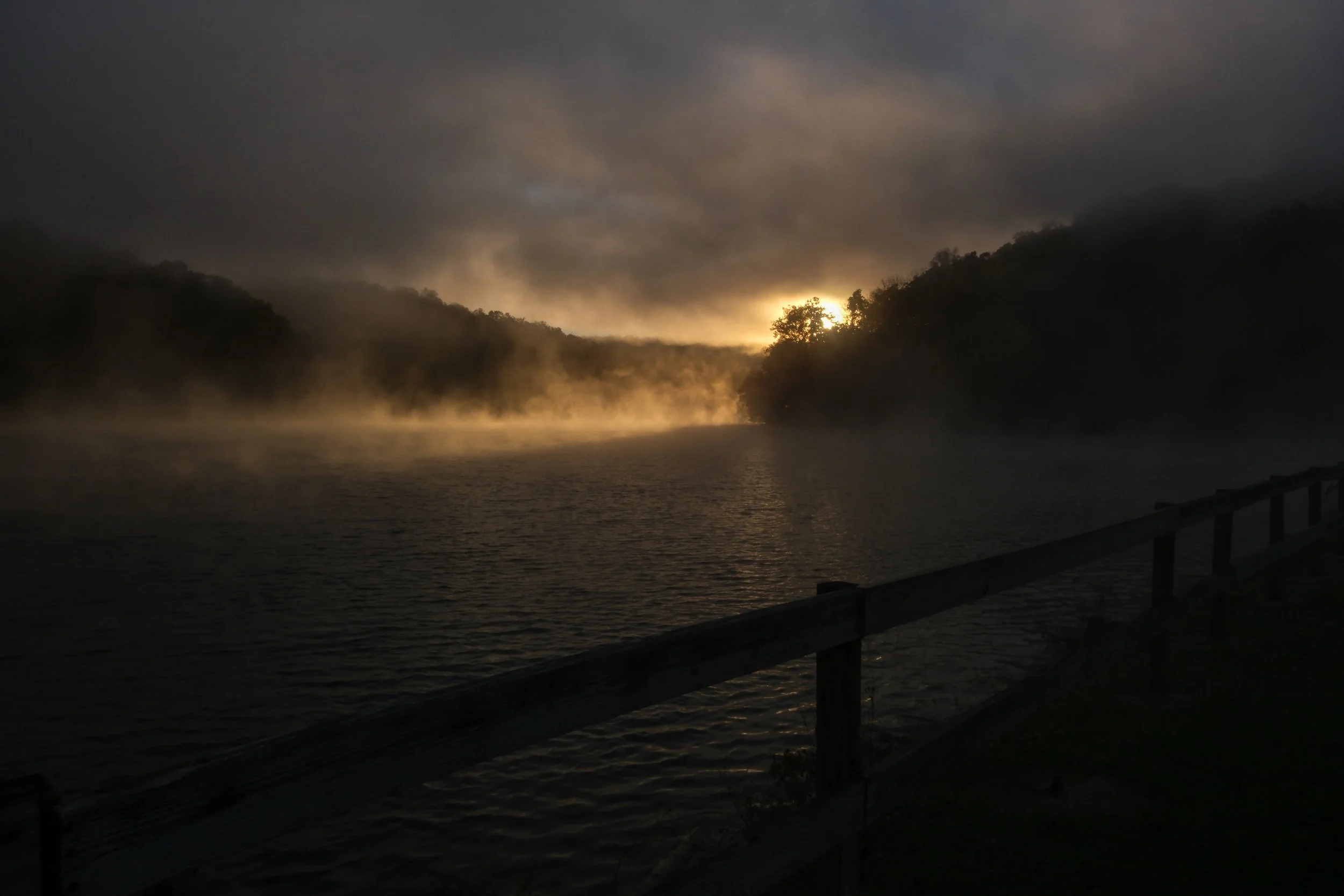 A river at dawn with mist rising and the sun peeking through cloudy skies, flanked by dark wooded hills, and a wooden railing in the foreground.