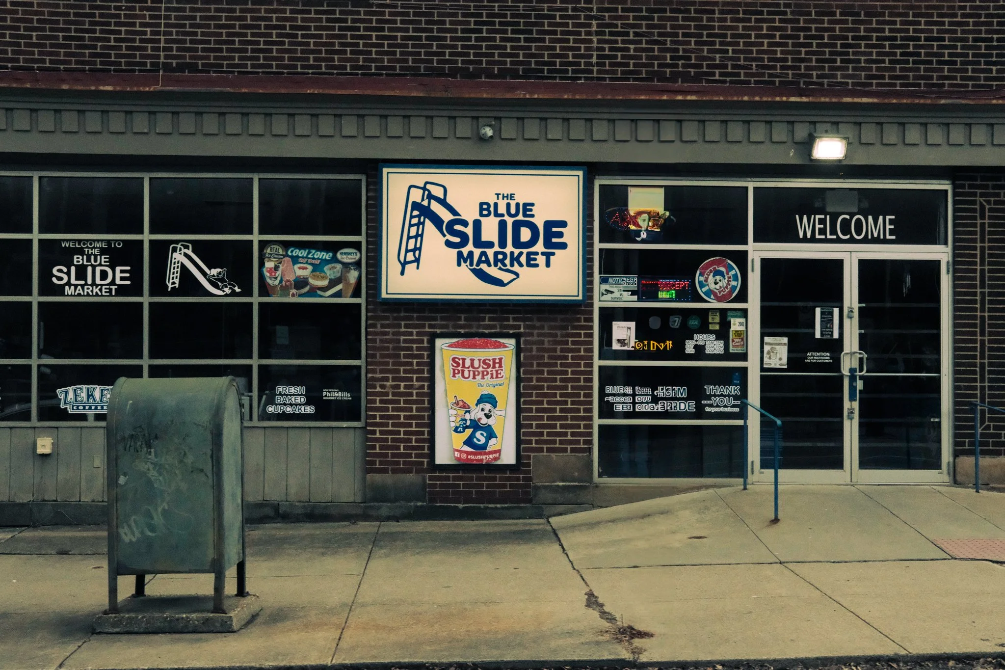 A storefront with signs reading 'The Blue Slide Market,' 'WELCOME,' and an advertisement for Slush Puppy with a cartoon dog mascot. The windows display ice cream and coffee ads, and there is a trash bin outside.