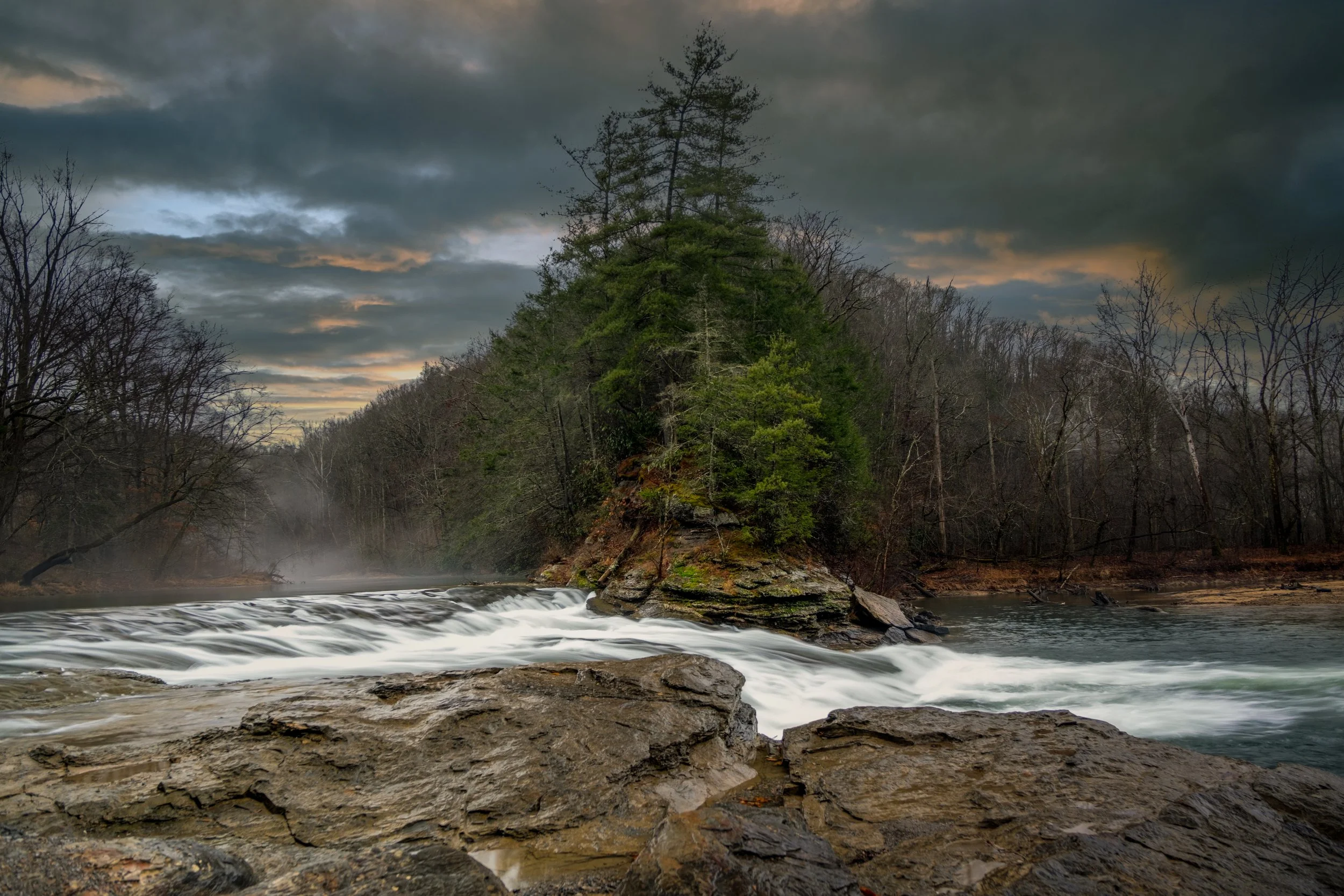A river with a small waterfall and an island with trees, under a cloudy sky.