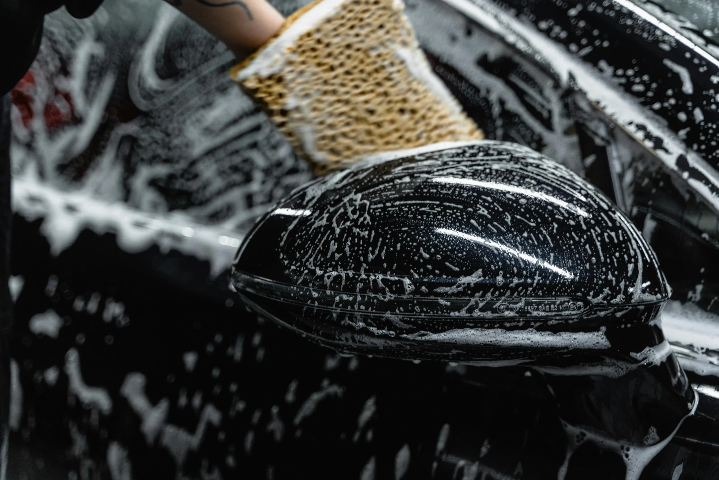 Close-up of a person washing a black car with a sponge and soap suds, focusing on the side mirror.