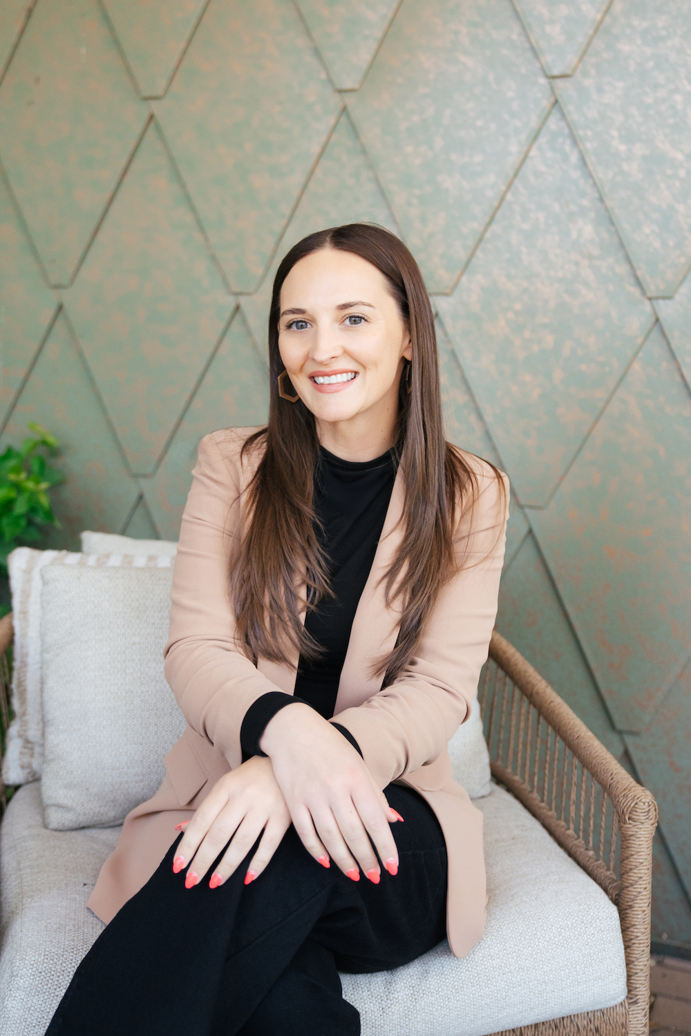 A woman with long brown hair and blue eyes smiling, sitting on a woven bench with beige cushions, in front of a green, geometric textured wall.