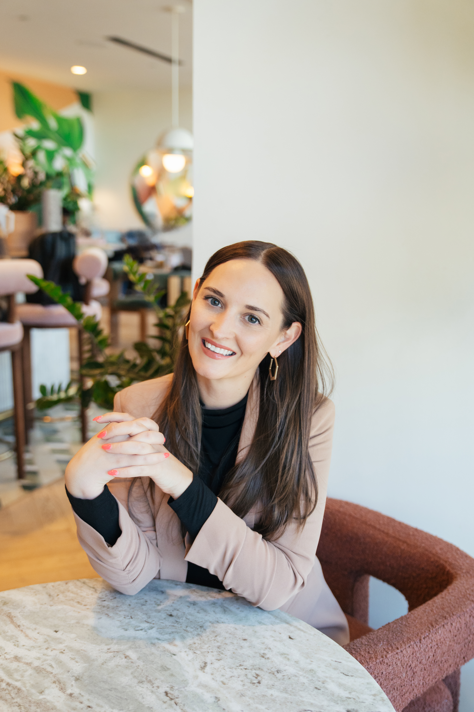 A woman with long brown hair and earrings sitting at a marble table in a modern, well-lit cafe or restaurant, smiling and looking at the camera.
