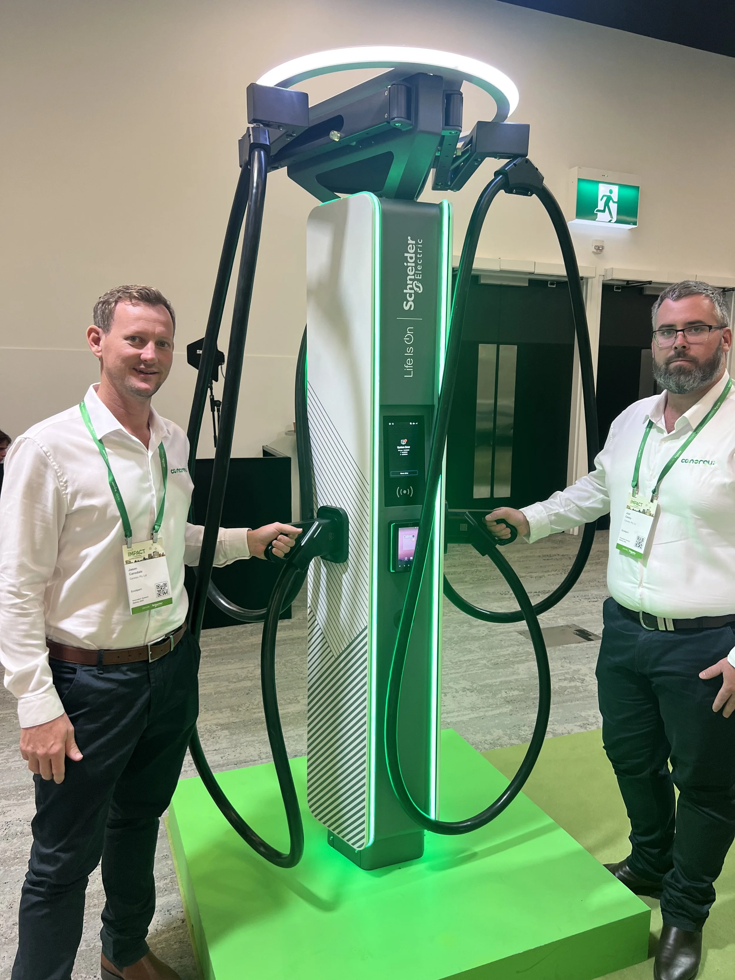 Jason and Joel standing next to a Schneider Electric EV charging station, featuring illuminated green lights, on a green platform. They are holding charging cables.
