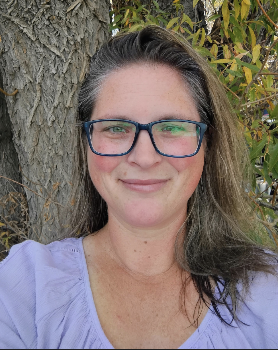 A woman with glasses and long hair smiling outdoors, with a large tree and green foliage in the background.