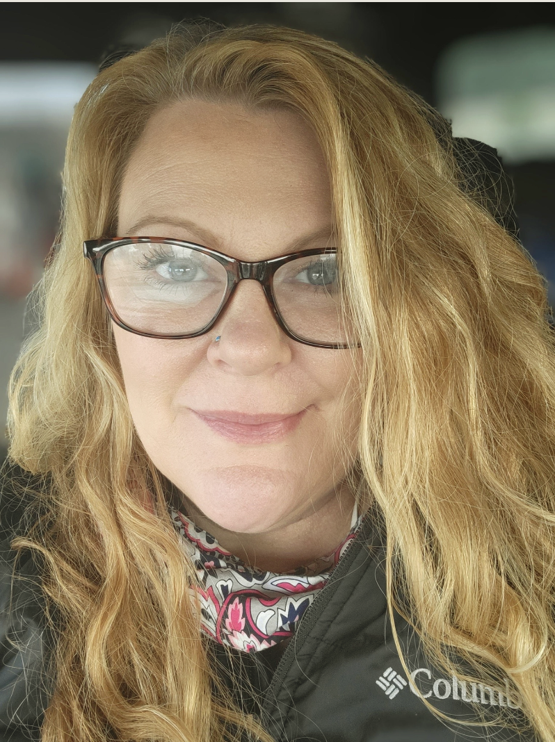 A woman with curly red hair wearing glasses, a black Columbia jacket, and a colorful neck gaiter, smiling at the camera inside a vehicle.
