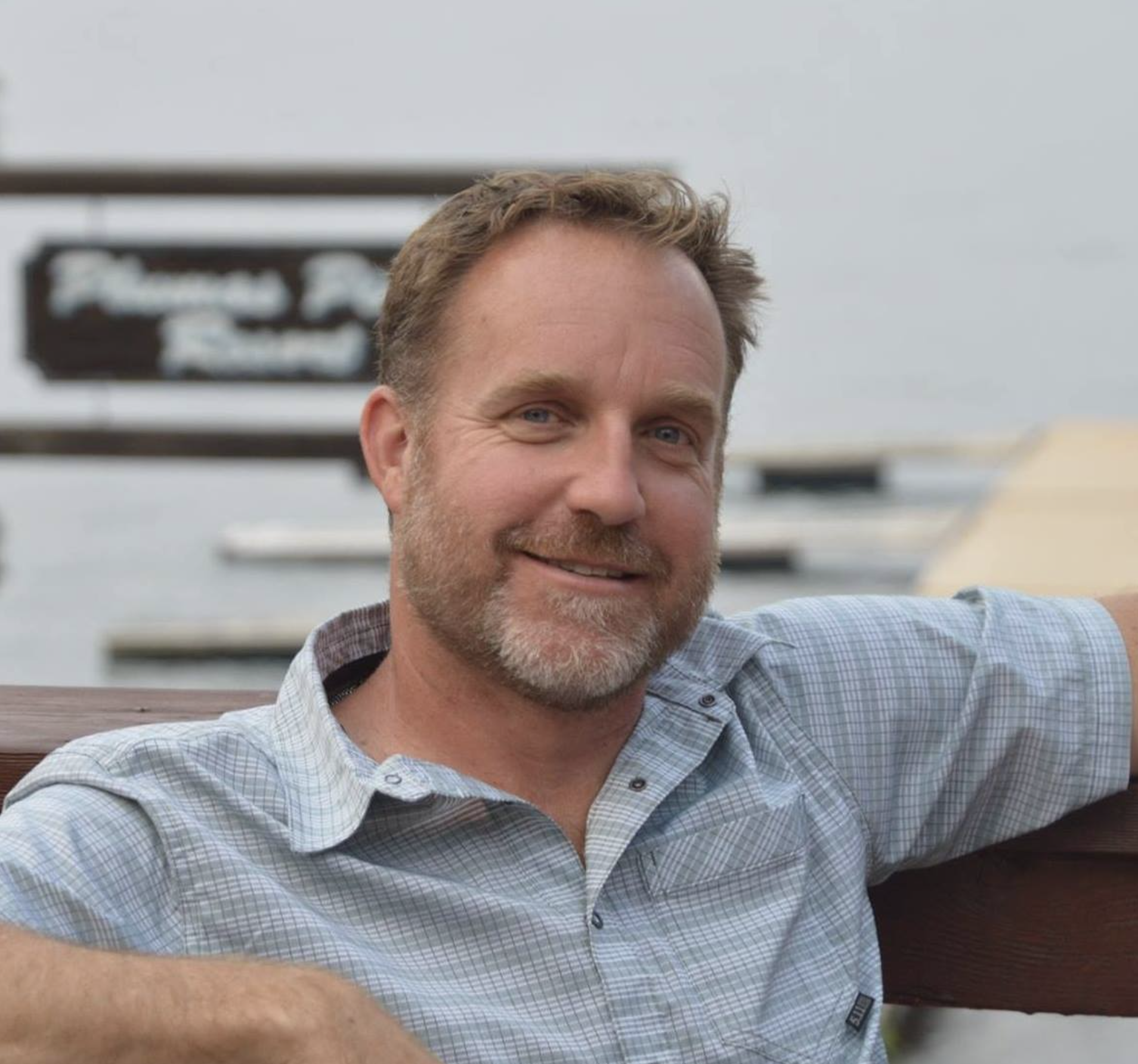 A man with short hair and a beard, wearing a light plaid shirt, smiling while sitting on a wooden bench outdoors.