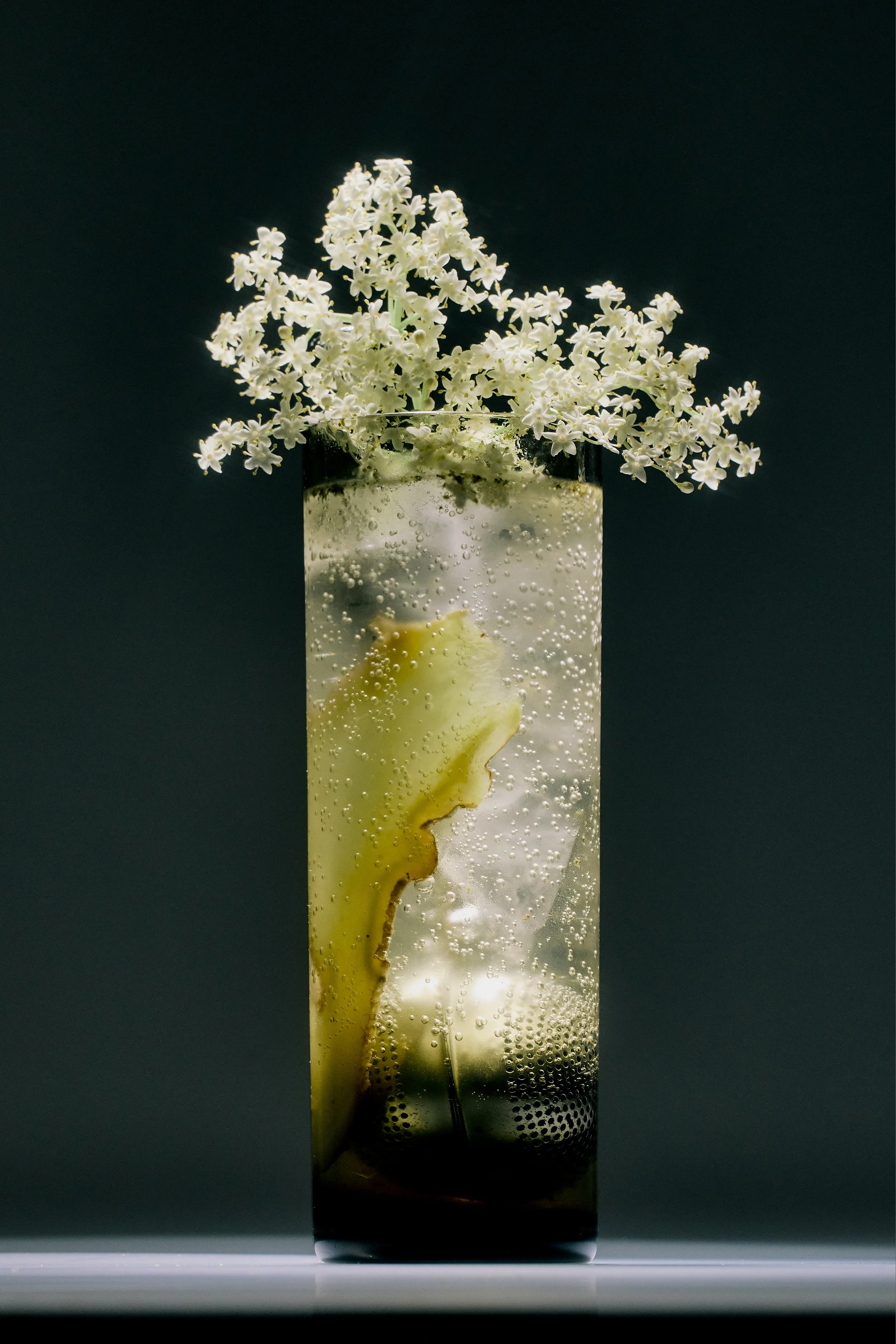 A tall glass filled with a fizzy drink garnished with elderflower blossoms and a slice of ginger, set against a dark background.