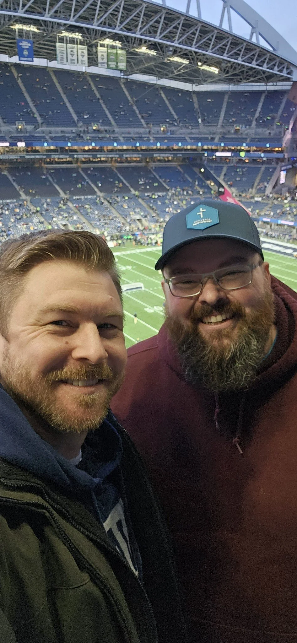 Two men smiling at a stadium with a football field in the background.