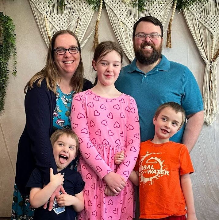 A family of five posing together in front of a decorative backdrop with macrame and greenery. The parents stand behind their three children, who are dressed casually. The atmosphere is cheerful and relaxed.