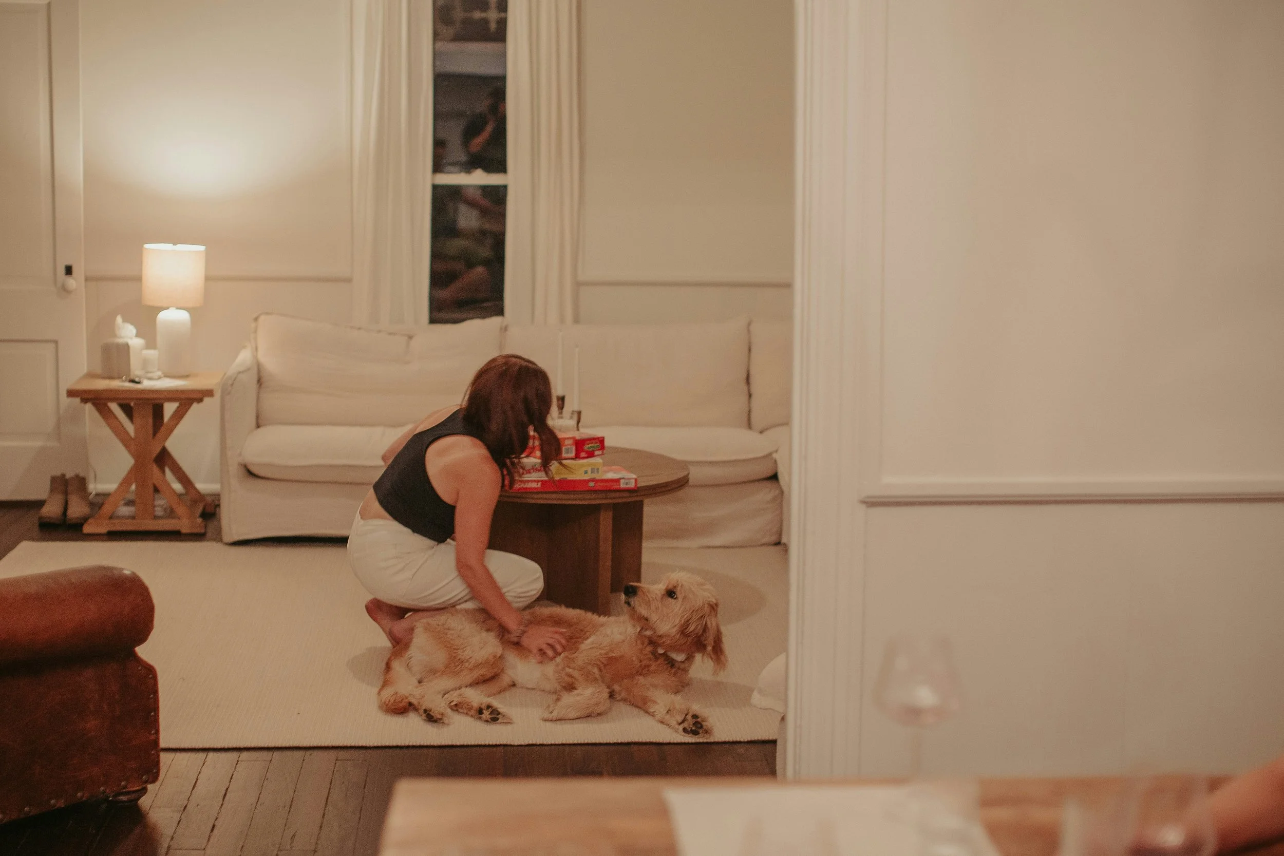 A woman kneeling on the floor petting a dog in a cozy living room with beige sofas, a wooden coffee table, and a side table with a lamp.