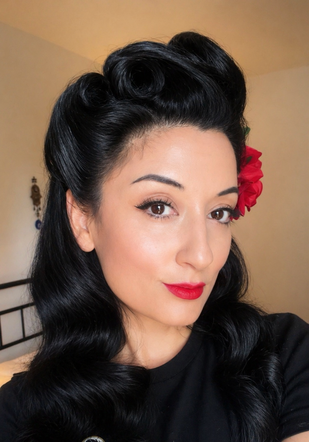 Close-up of a woman with vintage pin-up hairstyle, red lipstick, and a red flower in her hair. She has dark, wavy hair, brown eyes, and is wearing a black top.