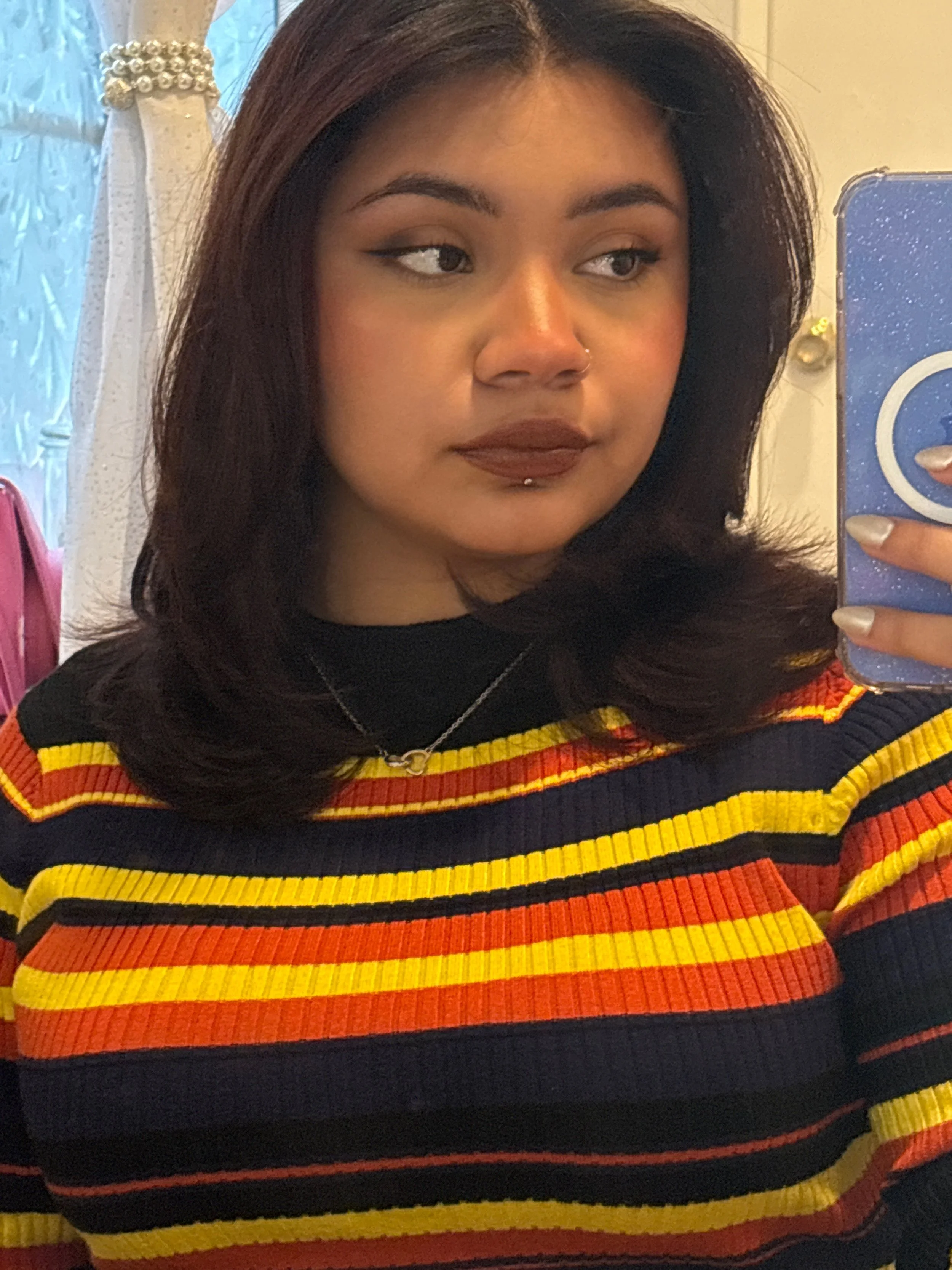 A young woman with shoulder-length dark hair taking a mirror selfie in a room with lace curtains. She has a nose and lip piercing, and is wearing a colorful striped sweater in red, yellow, black, and orange. She has makeup on, including dark lipstick and eyeliner.