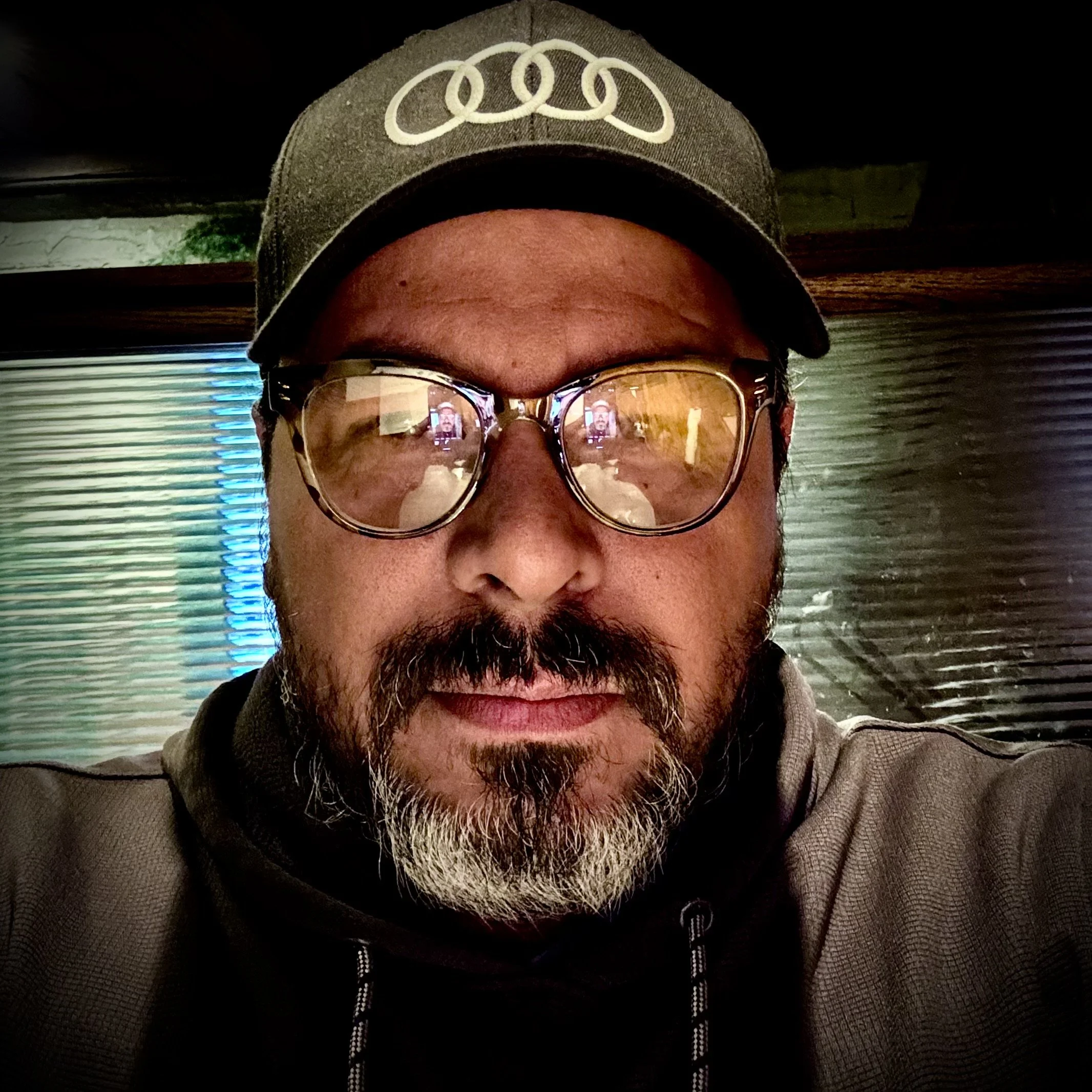 A close-up of a middle-aged man with curly black and gray hair, a full beard and mustache, wearing glasses and a white shirt.