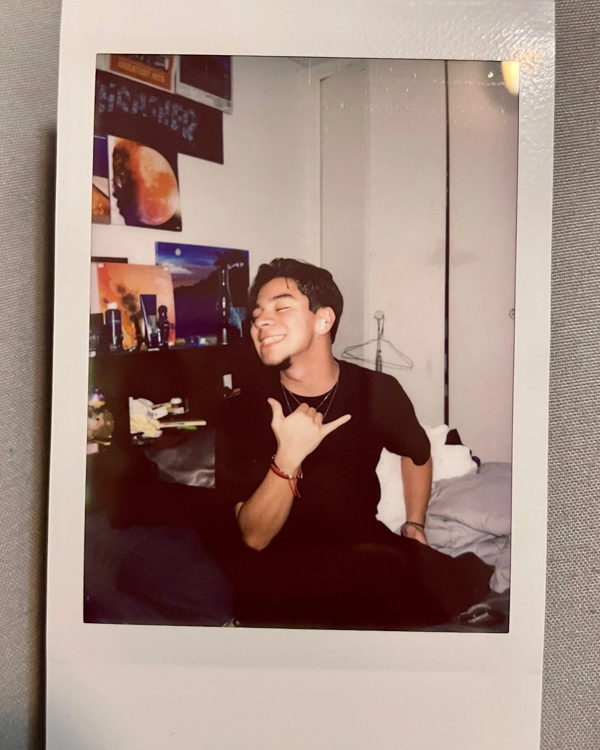 A young man with dark hair sitting on a bed, smiling with eyes closed, making a hand gesture. The room has posters and a computer on a desk behind him.
